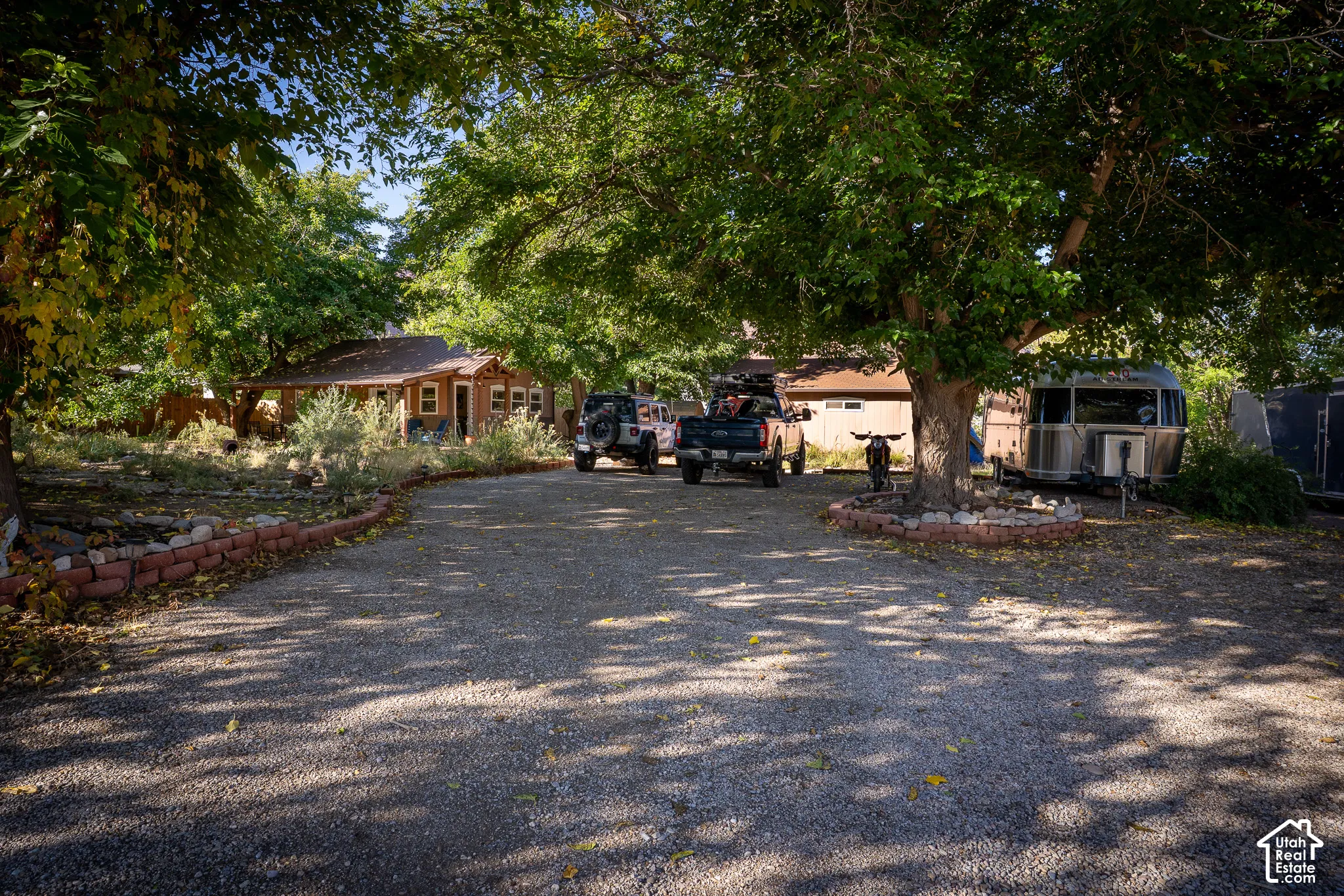 Gravel driveway featuring mature trees and a brick-bordered garden bed