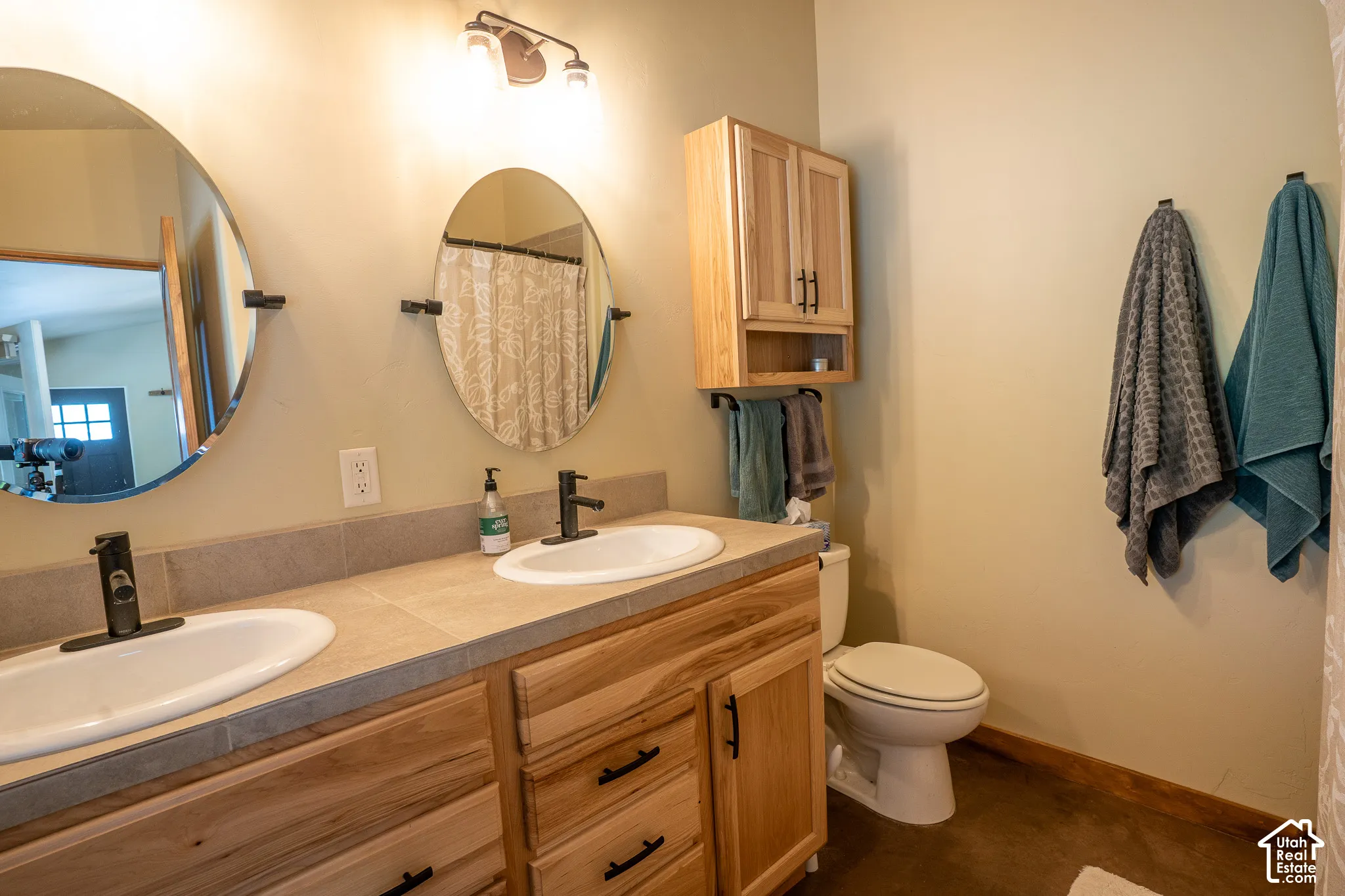 Dual-vanity cabinetry featuring natural wood finish, stone-tile countertop, oval vessel sinks, matte black fixtures, and wall-mounted wooden storage cabinet