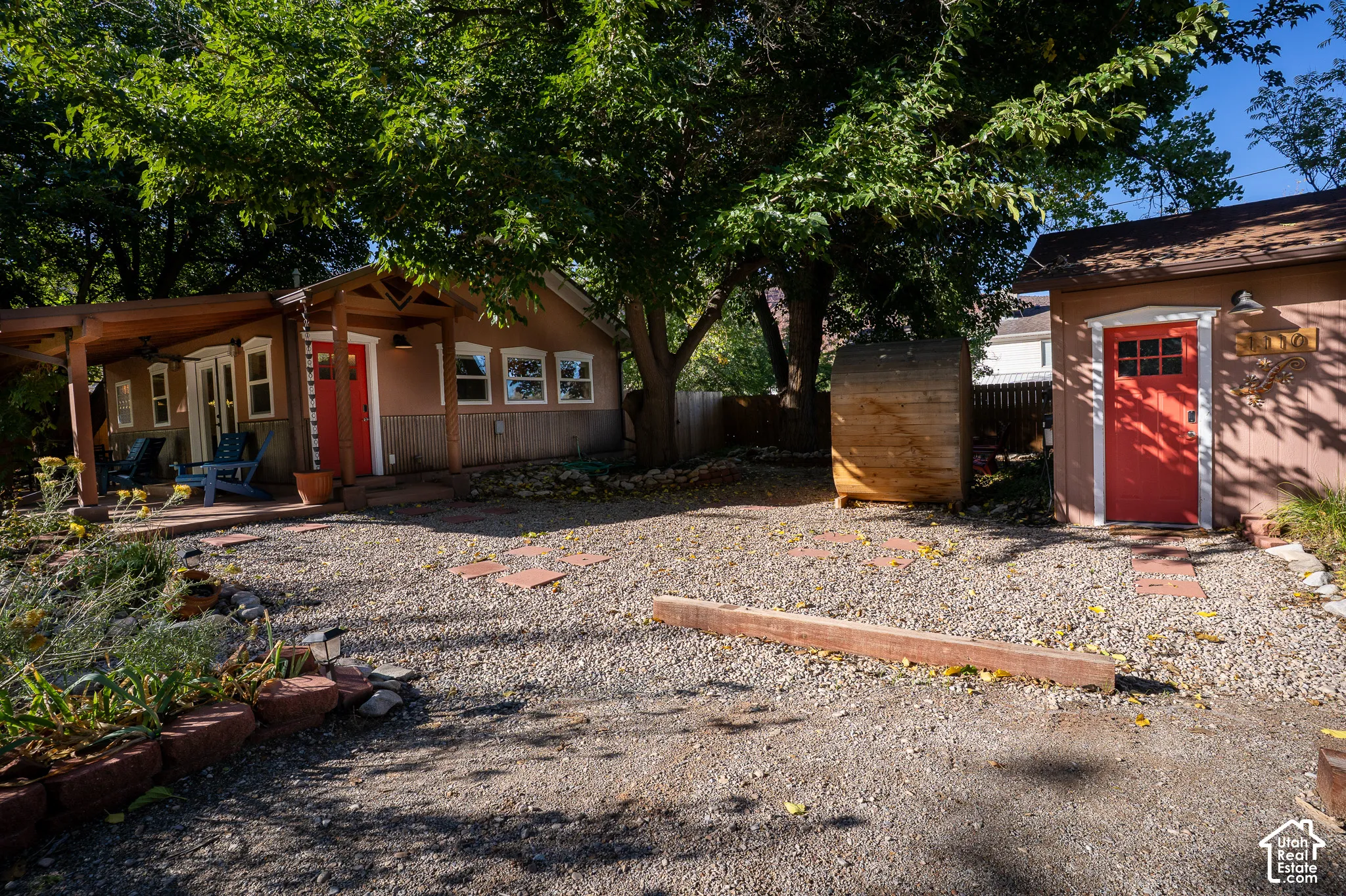 Gravel courtyard featuring a covered porch, detached storage structure with a red entry door, and mature canopy trees