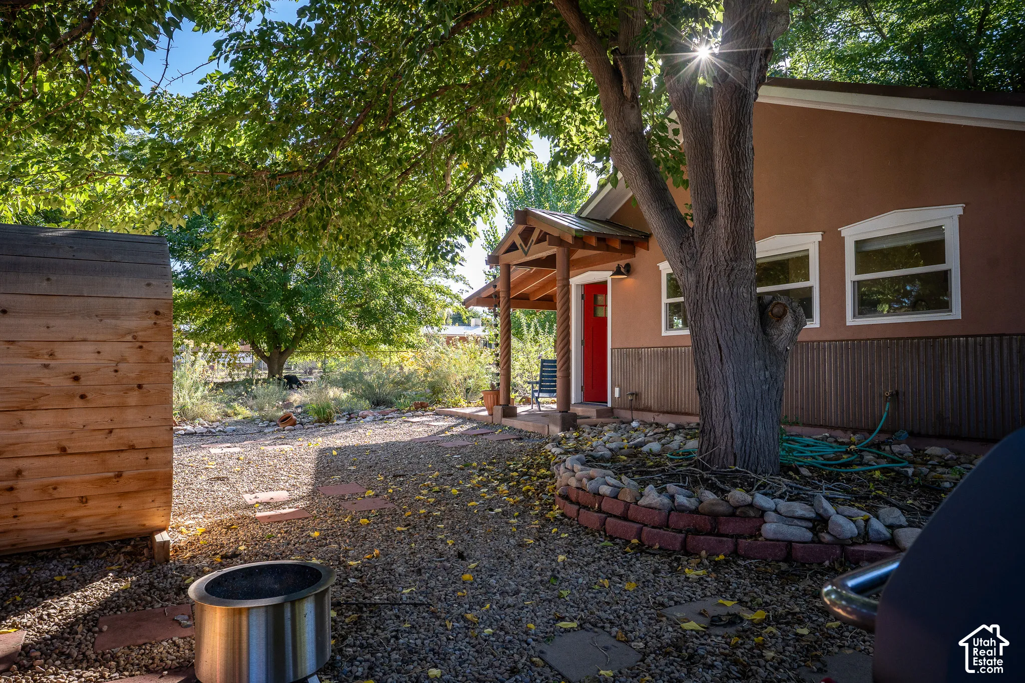 Exterior entrance featuring a covered porch with timber columns, stucco and vertical wood siding, a stone-bordered tree planter, and a gravel grounds area