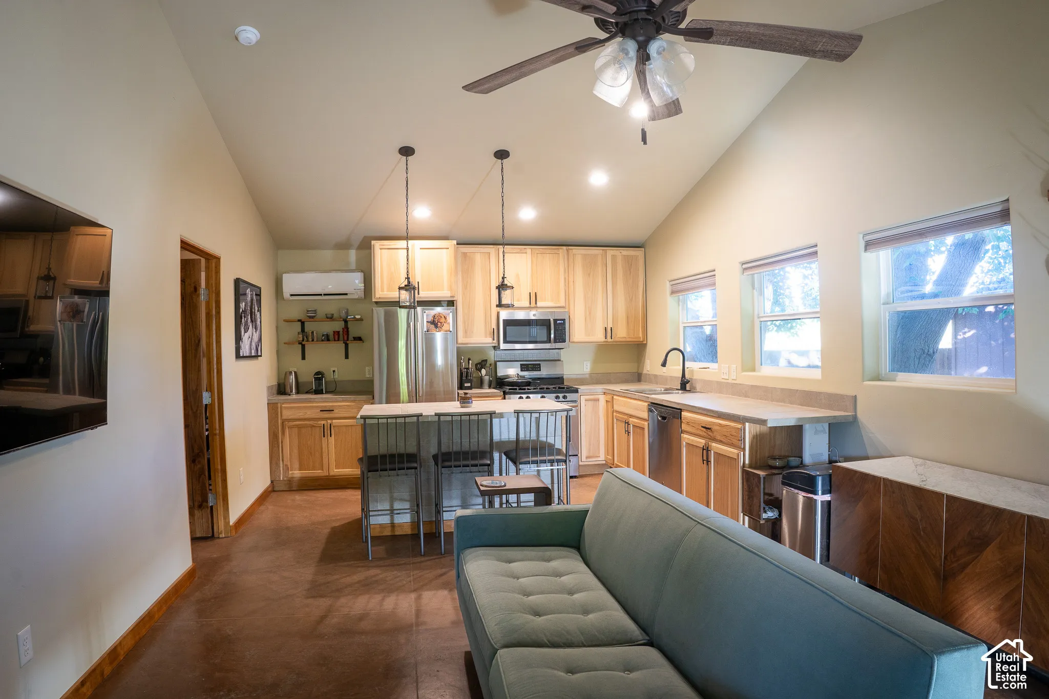 Vaulted ceiling with an integrated multi-blade ceiling fan, natural wood-finish cabinetry, central kitchen island, stone-finish countertops, and polished concrete flooring