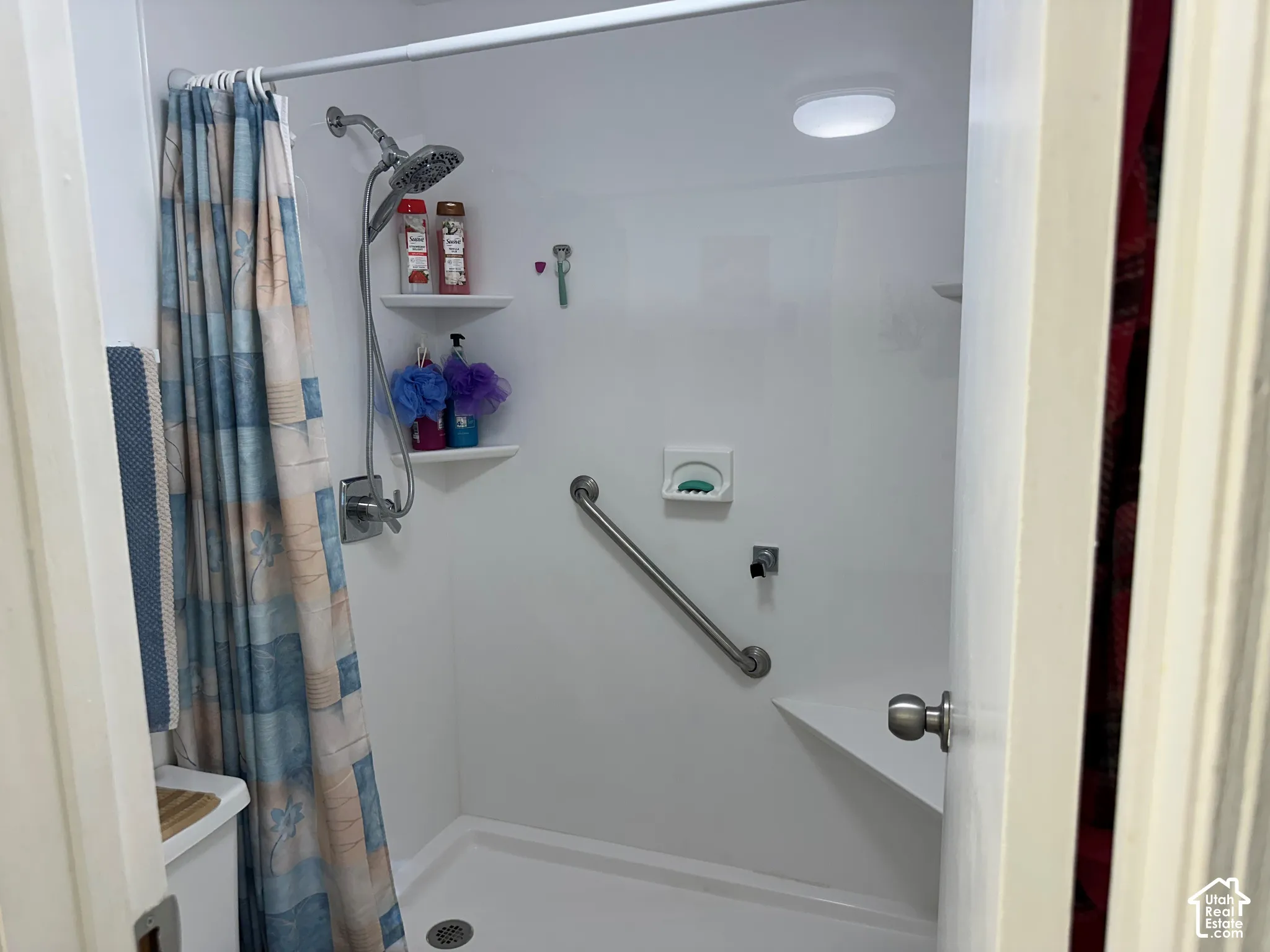 Shower stall featuring white wall panels, a handheld shower fixture, integrated shelving, a built-in corner seat, and a wall-mounted grab bar