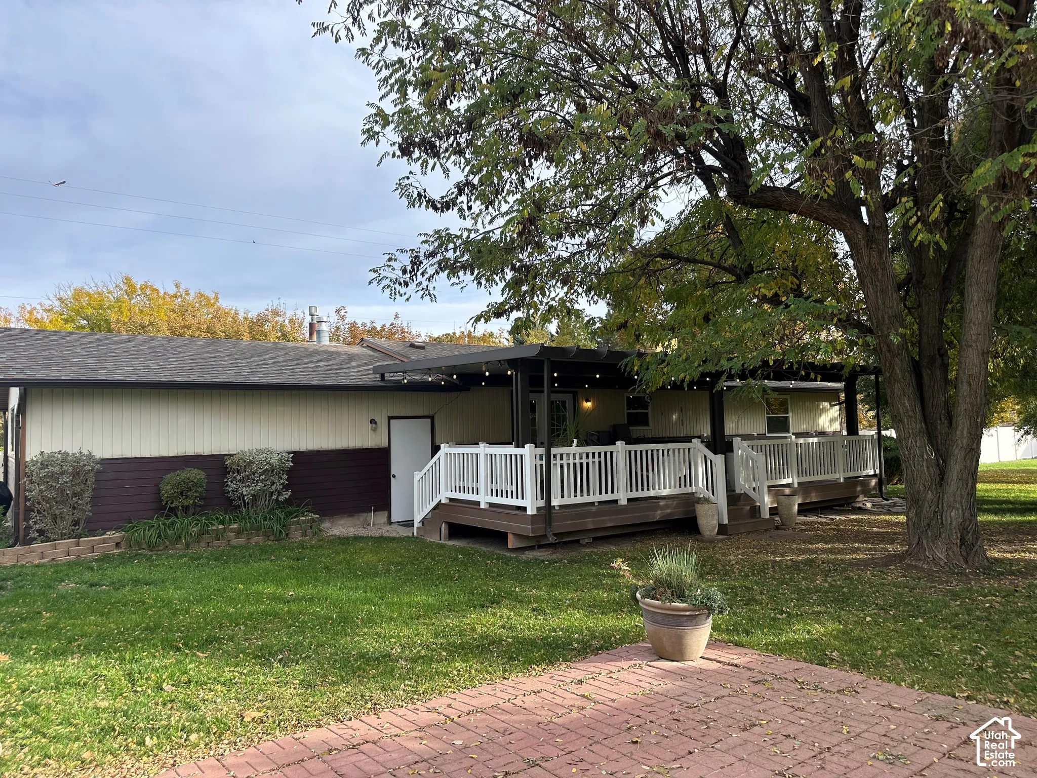 Rear exterior featuring a covered deck, white railings, brick paver patio, and mature tree canopy