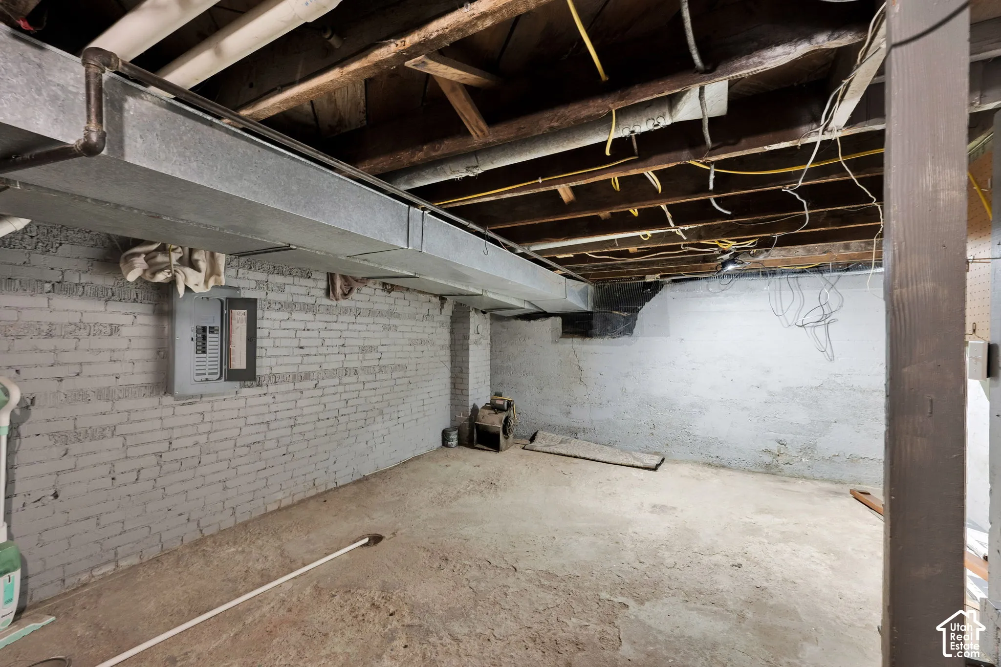 Unfinished basement area featuring painted masonry walls, exposed wood ceiling joists, metal HVAC ductwork, and a wall-mounted electrical panel