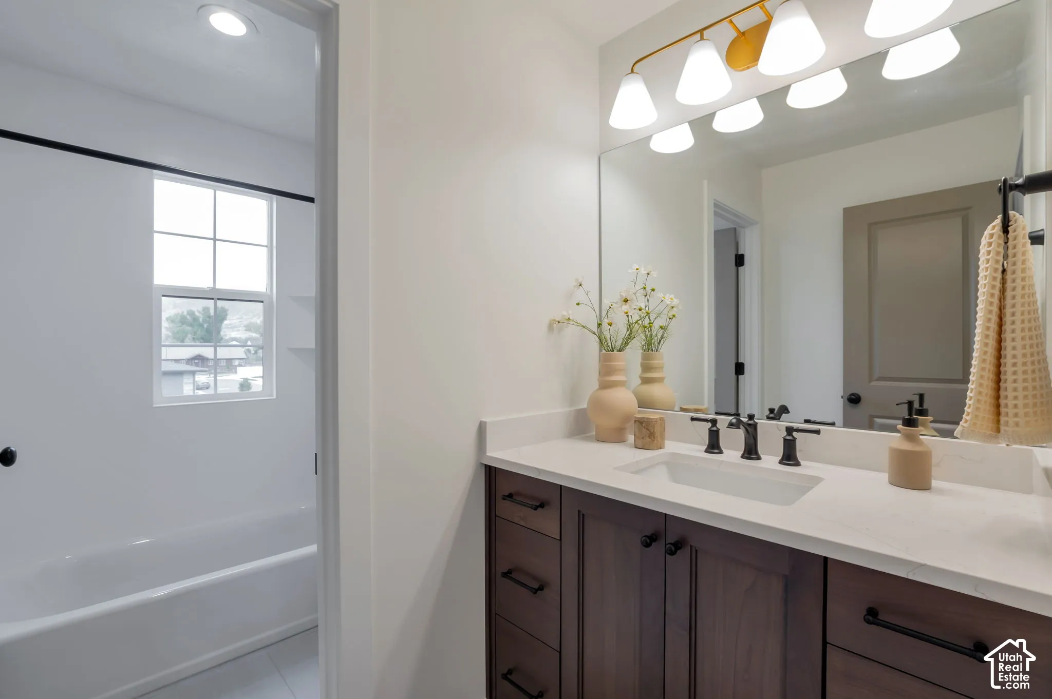 Bathroom featuring a vanity with wood-finish cabinetry, light-toned stone-surface countertop, rectangular undermount sink, matte black hardware, and an expansive wall-mounted mirror with multi-bulb overhead lighting