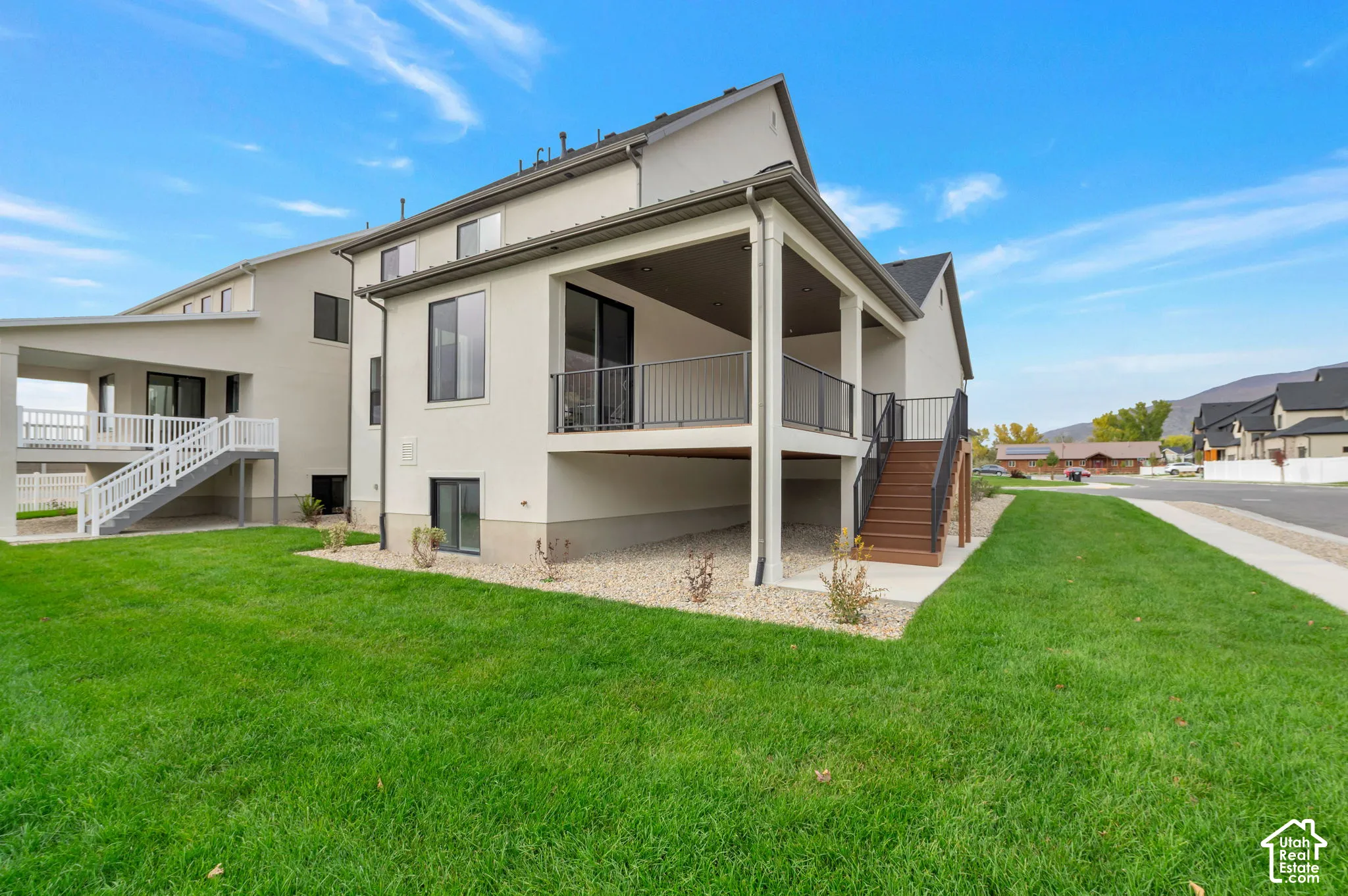 Two-story exterior featuring light-colored stucco siding, a covered rear deck with black metal railing, and wood-finish stairs leading to the lawn