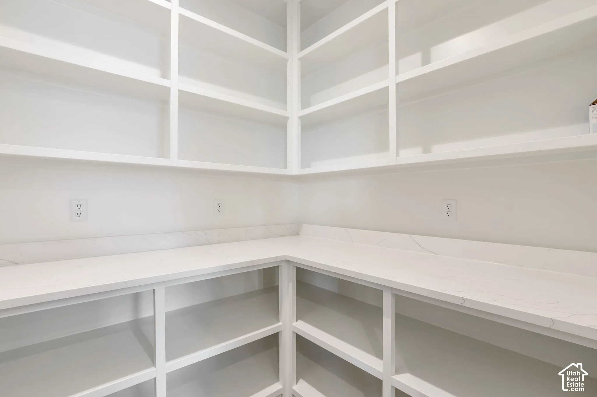 Walk-in storage area featuring custom white built-in shelving, stone-composite countertops with matching backsplash, and integrated electrical outlets