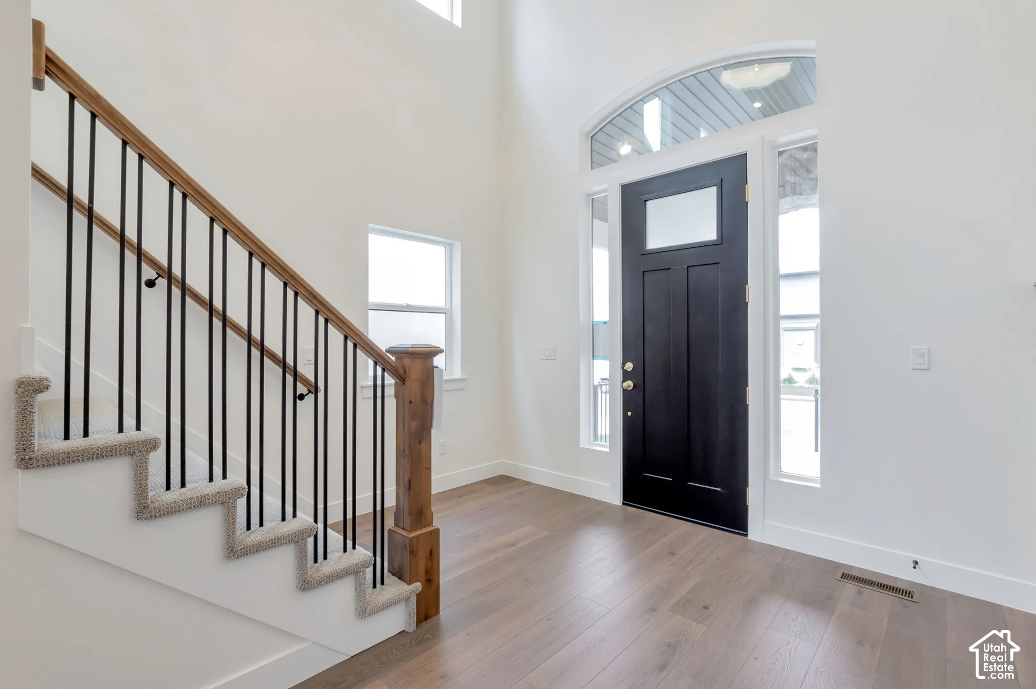 Entryway featuring vaulted ceilings, wide-plank wood-finish flooring, a solid black front door with a transom window, and a staircase with a stained wood handrail and iron balusters