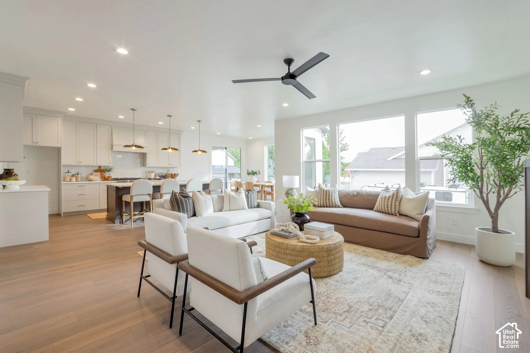 Open-concept area featuring light-toned wood-finish flooring, recessed lighting, a modern ceiling fan, large windows, and a kitchen equipped with a center island and pendant lighting