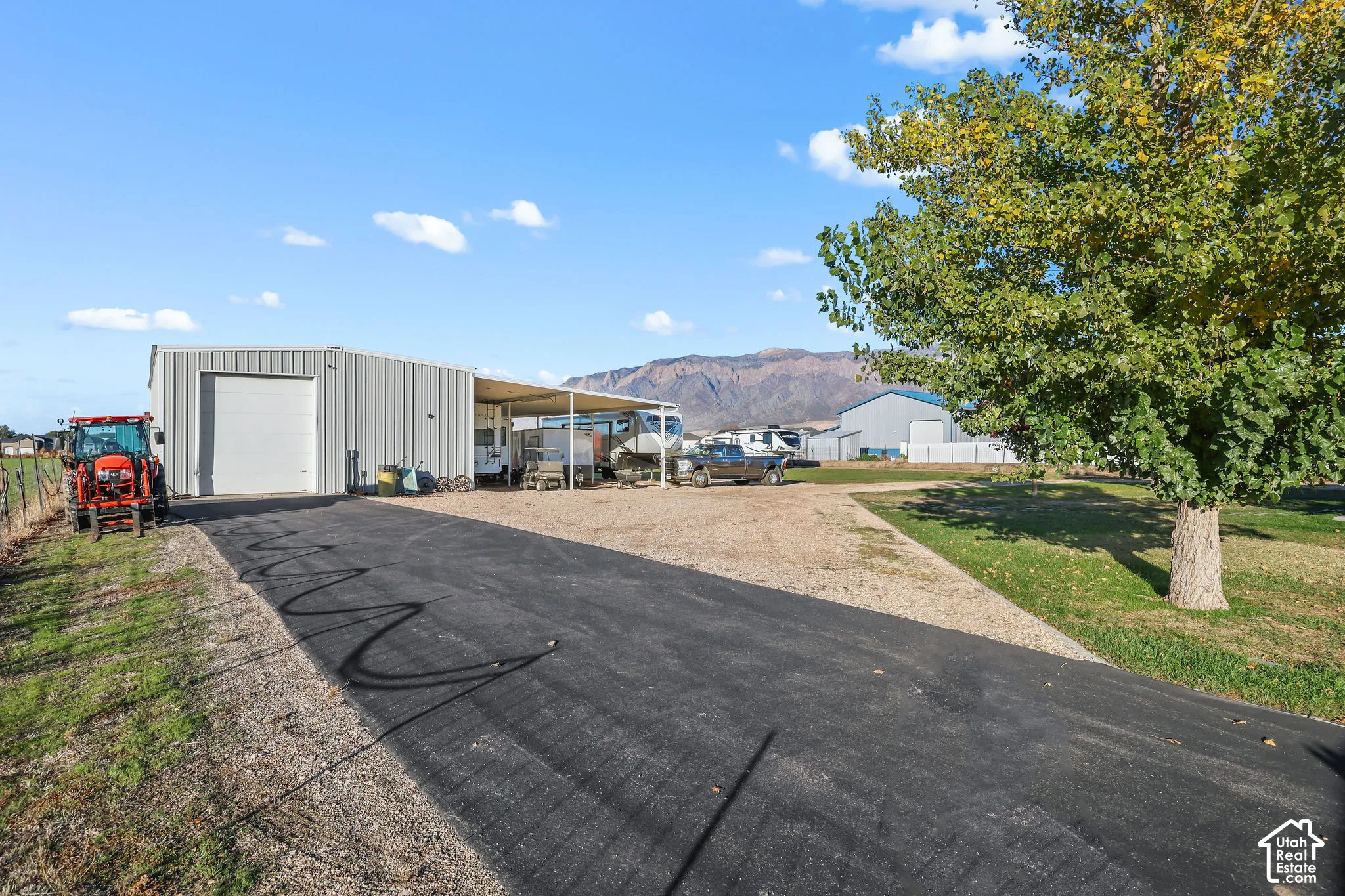 Metal-sided workshop featuring a high-clearance roll-up door, an attached covered carport, asphalt driveway, and mountain backdrop