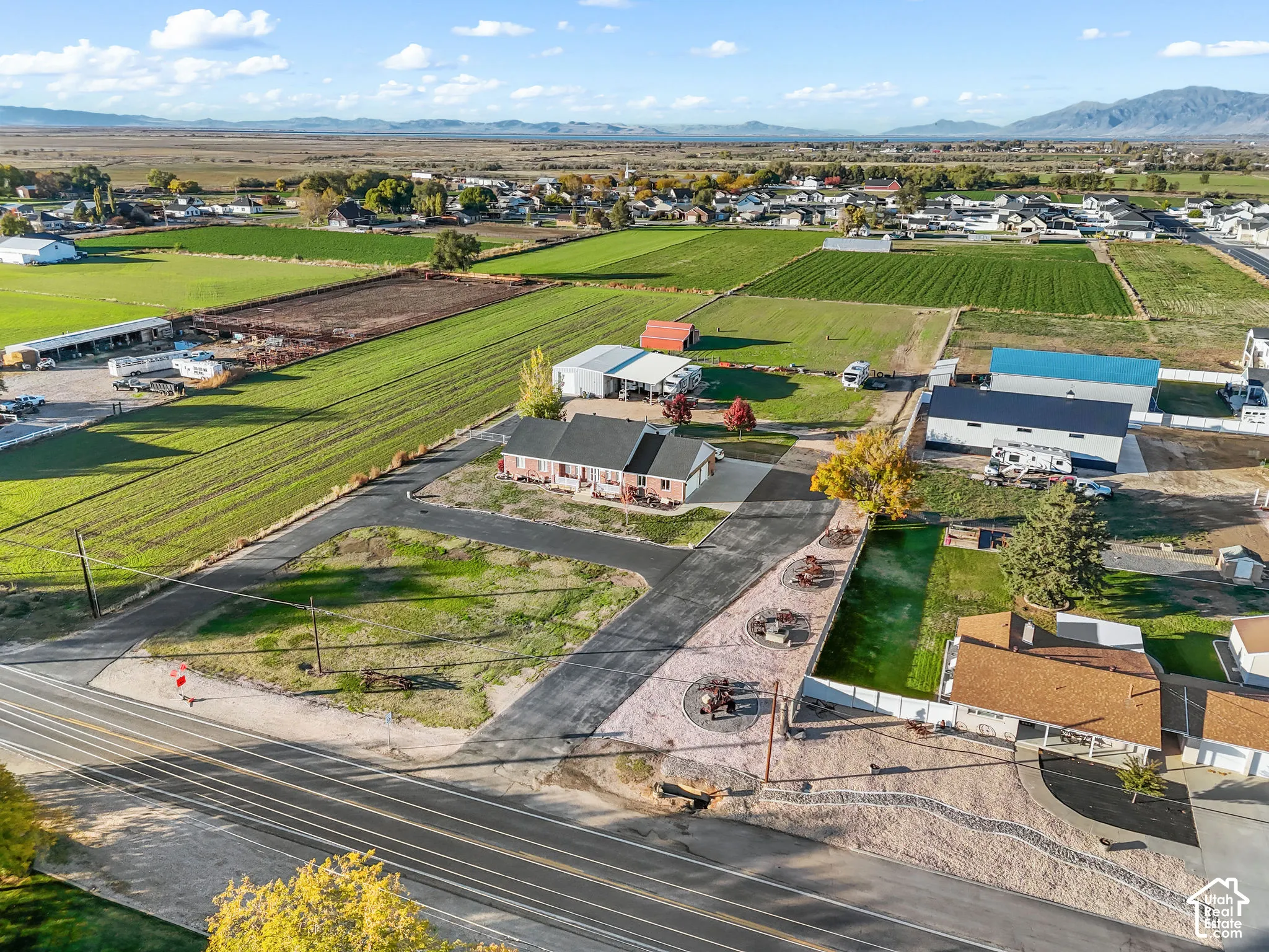 Aerial perspective featuring an asphalt driveway, brick residence, detached metal outbuilding, mountain backdrop, and expansive open acreage