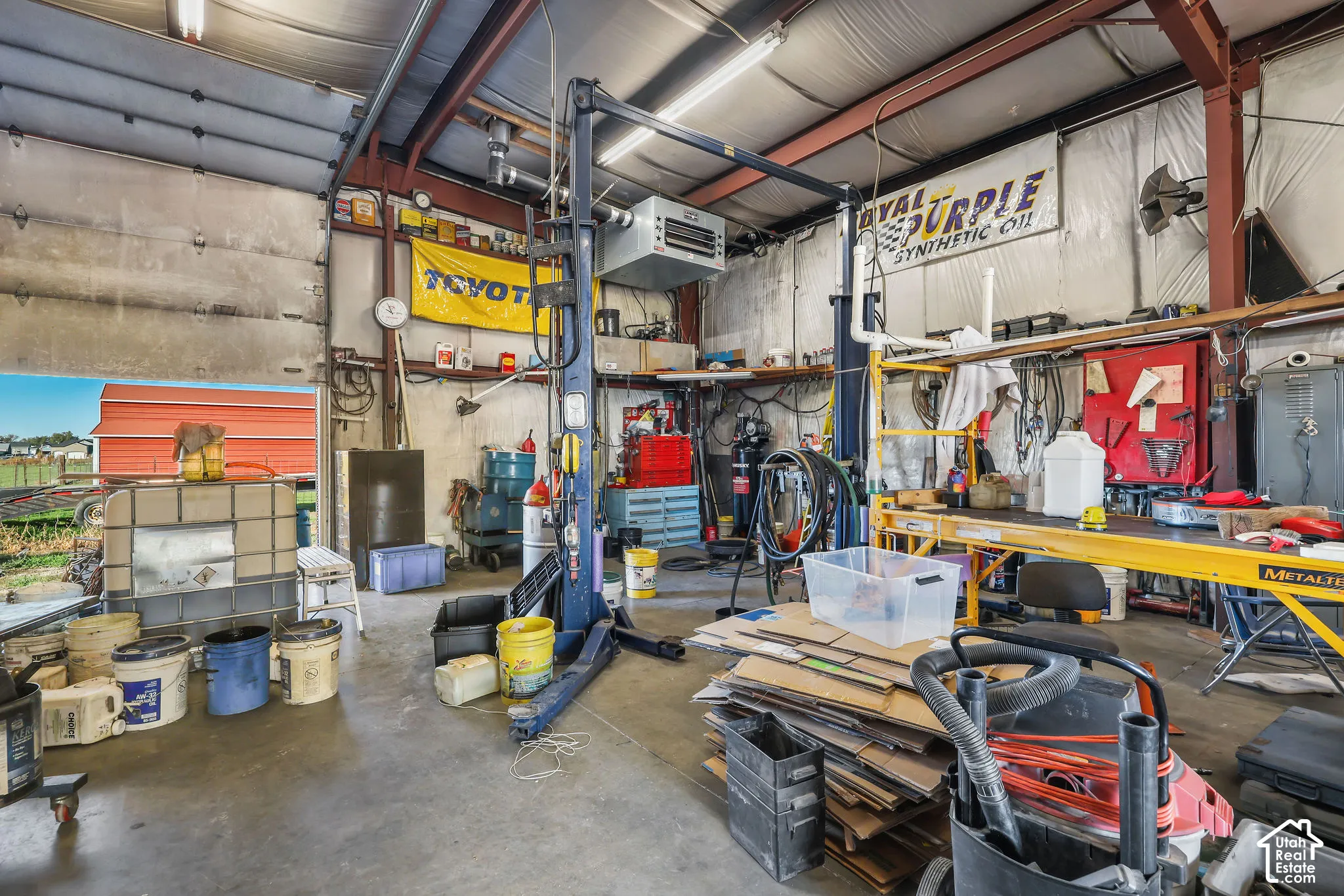 Expansive workshop featuring exposed steel framing, high-clearance overhead garage door, concrete flooring, built-in hydraulic vehicle lift, and overhead industrial heating unit