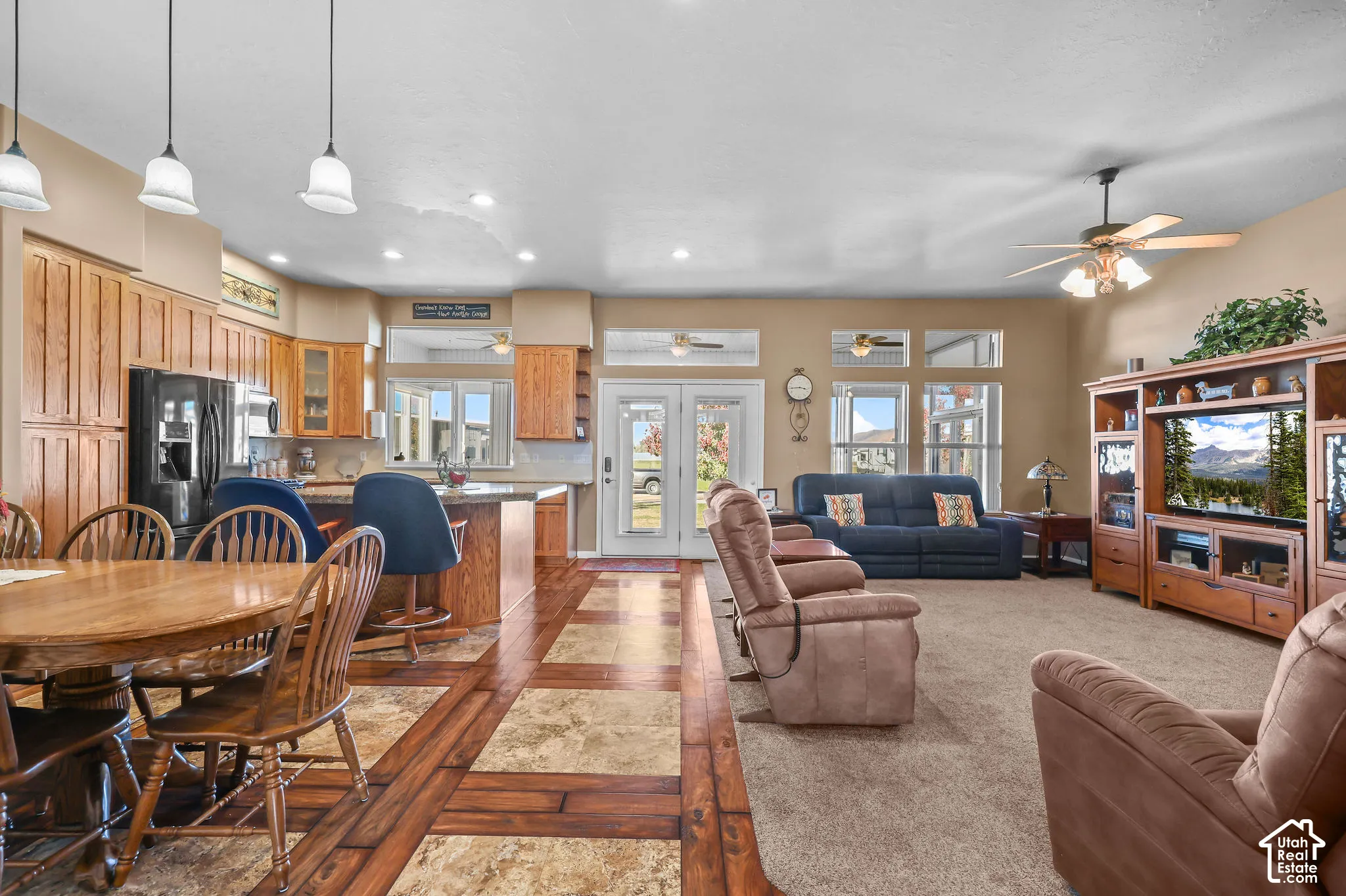 Open-concept interior featuring light wood cabinetry, multi-texture flooring with inset stone-finish tiles and wood-finish borders, recessed lighting, double French doors, and wooden entertainment cabinetry