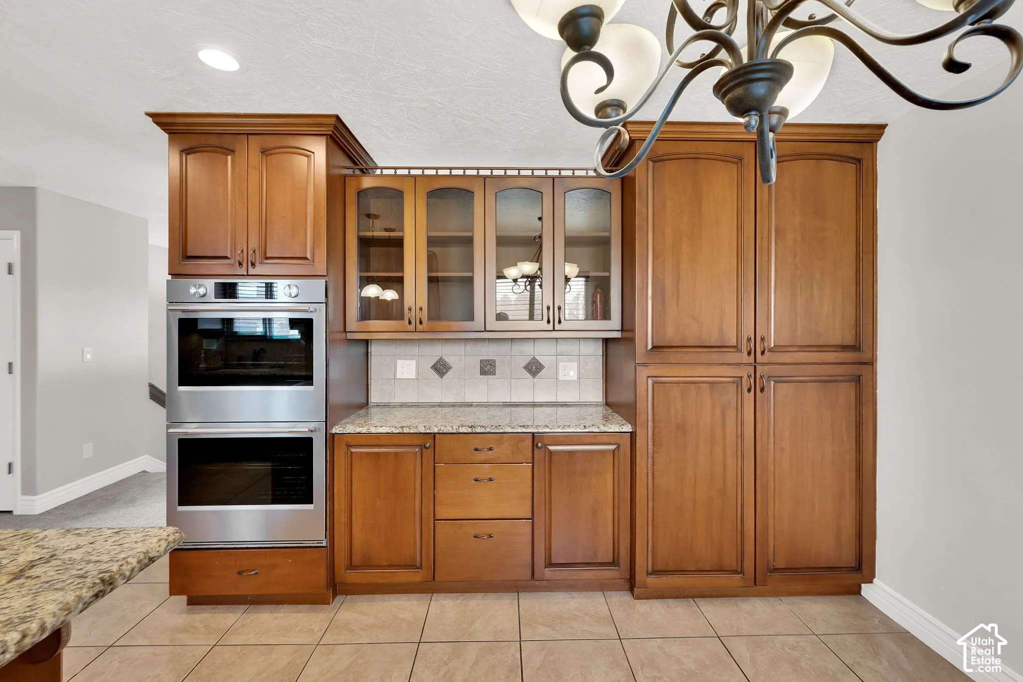 Custom wood cabinetry featuring glass-front display panels, stainless steel double wall ovens, granite countertops, tile backsplash with decorative diamond inlay, and ceramic tile flooring