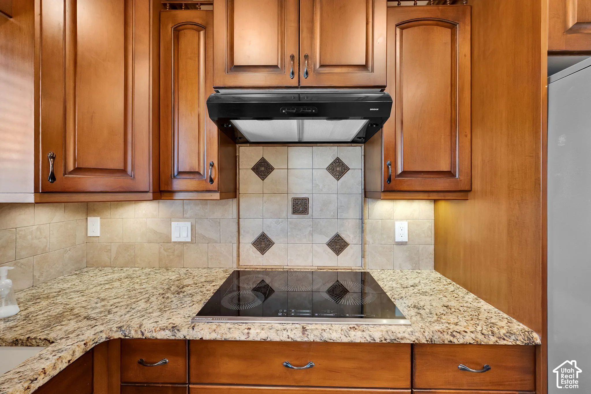 Kitchen featuring wood cabinetry, stone-finish countertops, black electric cooktop, matching range hood, and decorative tile backsplash with inlay accents