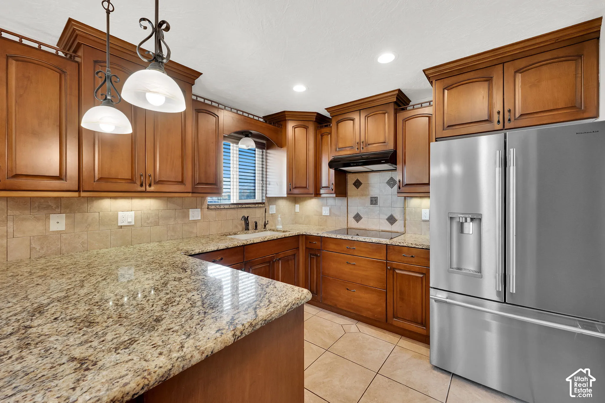 Kitchen featuring stained wood cabinetry, stone countertops, tiled backsplash, stainless steel refrigerator, and recessed lighting