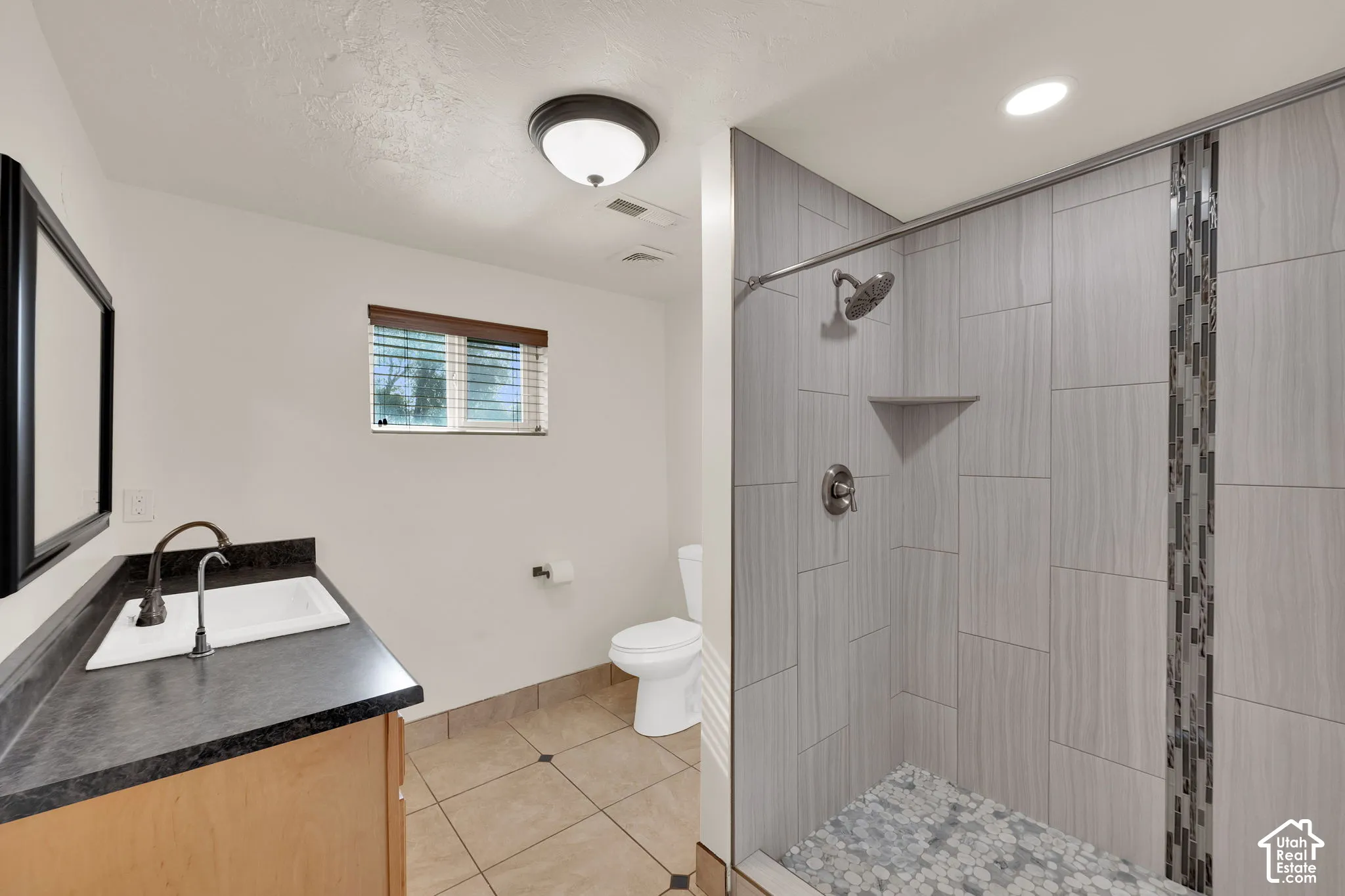 Bathroom featuring wood-finish cabinetry, dark solid-surface countertop with an integrated basin, tile flooring, and a walk-in shower with vertical decorative glass tile inlay and pebble-style floor tiling