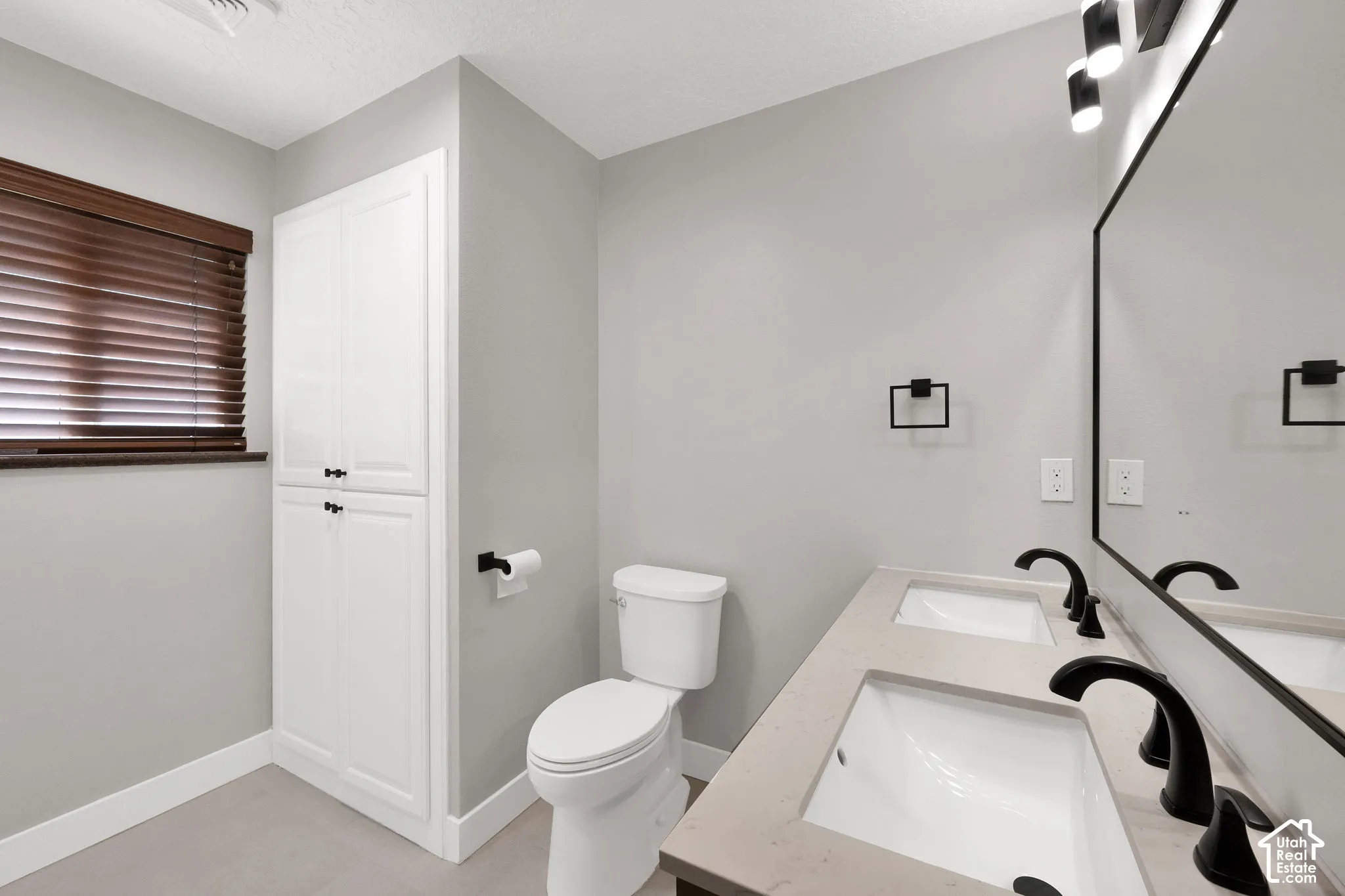 Bathroom featuring a dual-sink vanity with a solid-surface countertop, matte black fixtures, built-in cabinetry, and horizontal window blinds