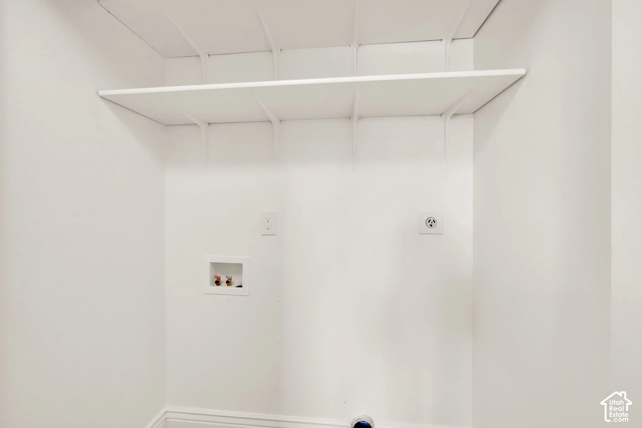 Utility area featuring white walls, overhead shelving supported by wall-mounted brackets, a recessed water supply box, and dedicated electrical outlets
