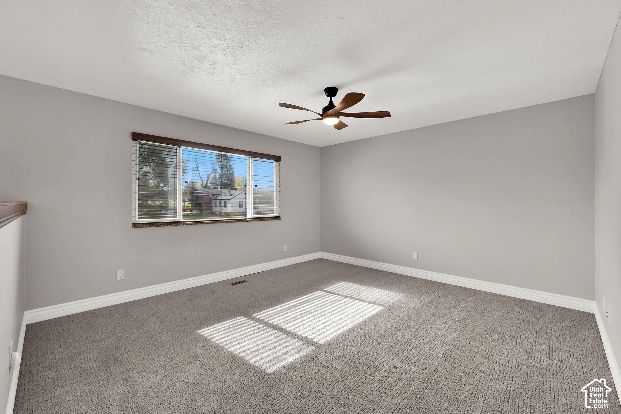 Neutral-toned interior space featuring neutral carpeting, a multi-blade ceiling fan with integrated lighting, and a large window with horizontal blinds