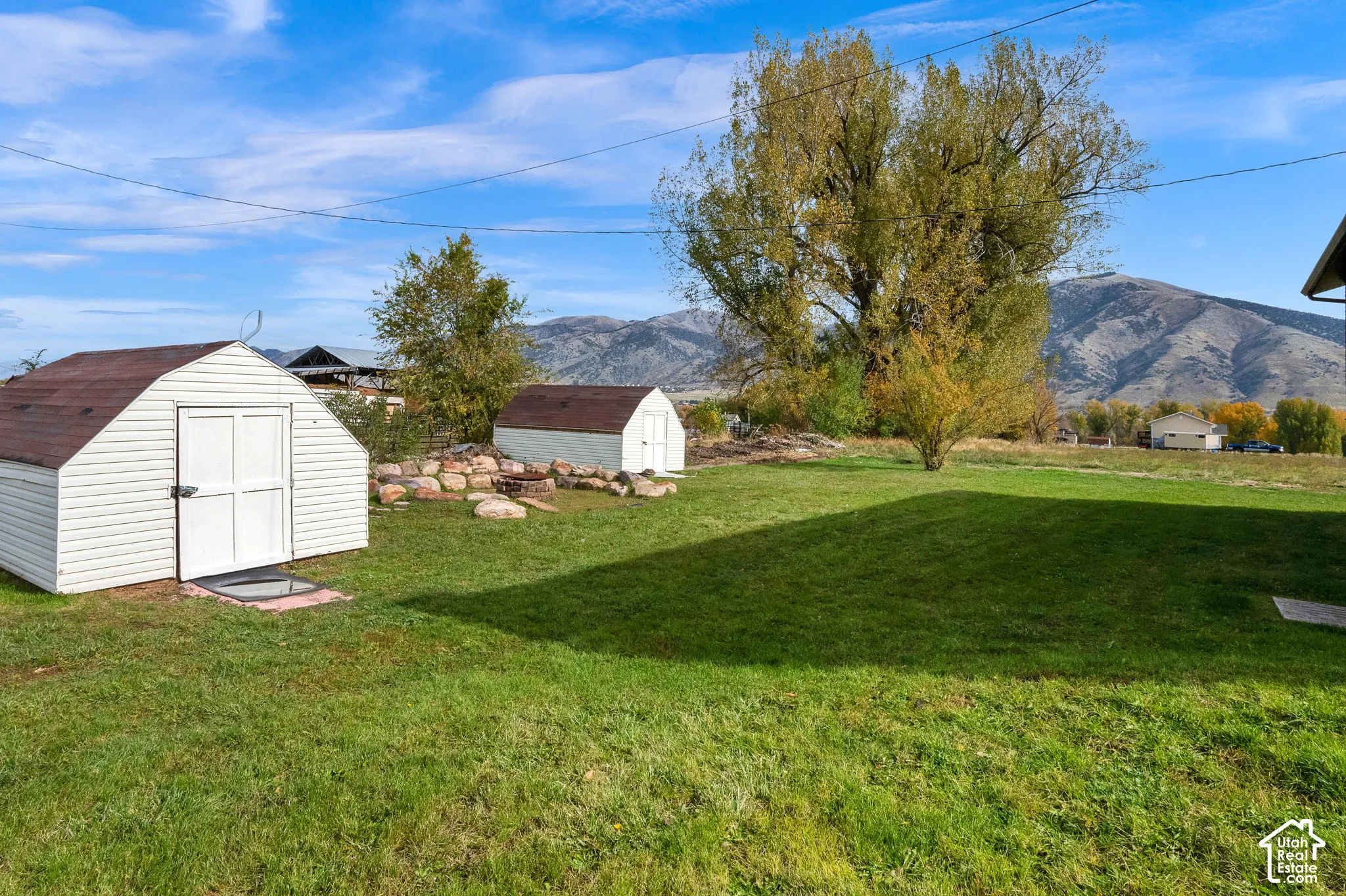 Expansive backyard featuring two storage sheds, a stone-lined fire pit area, mature trees, and a mountain backdrop