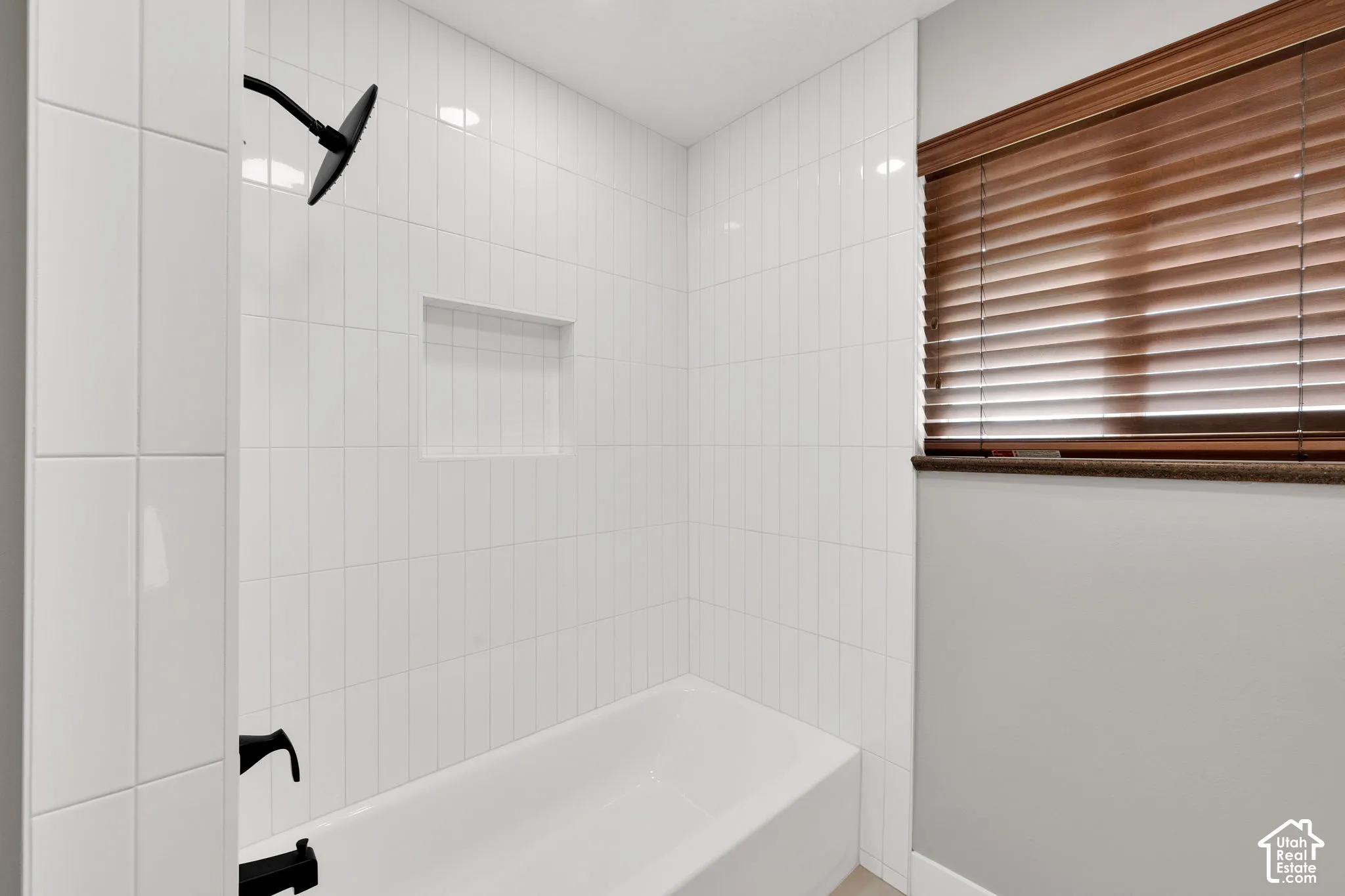 Shower and bathtub combination featuring floor-to-ceiling white rectangular wall tile, a recessed niche, matte black fixtures, and a window with wood-finish horizontal blinds