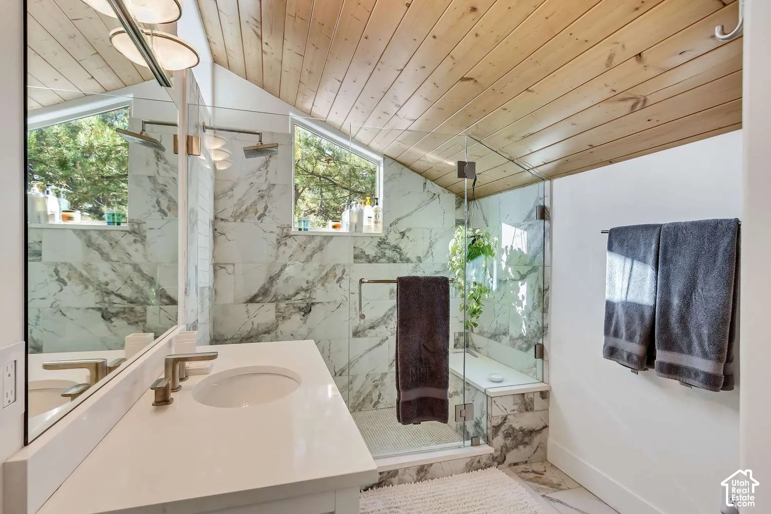 Bathroom featuring a tongue-and-groove wood-paneled sloped ceiling, stone-tiled shower enclosure with built-in bench, frameless glass shower door, and a white vanity with an integrated basin