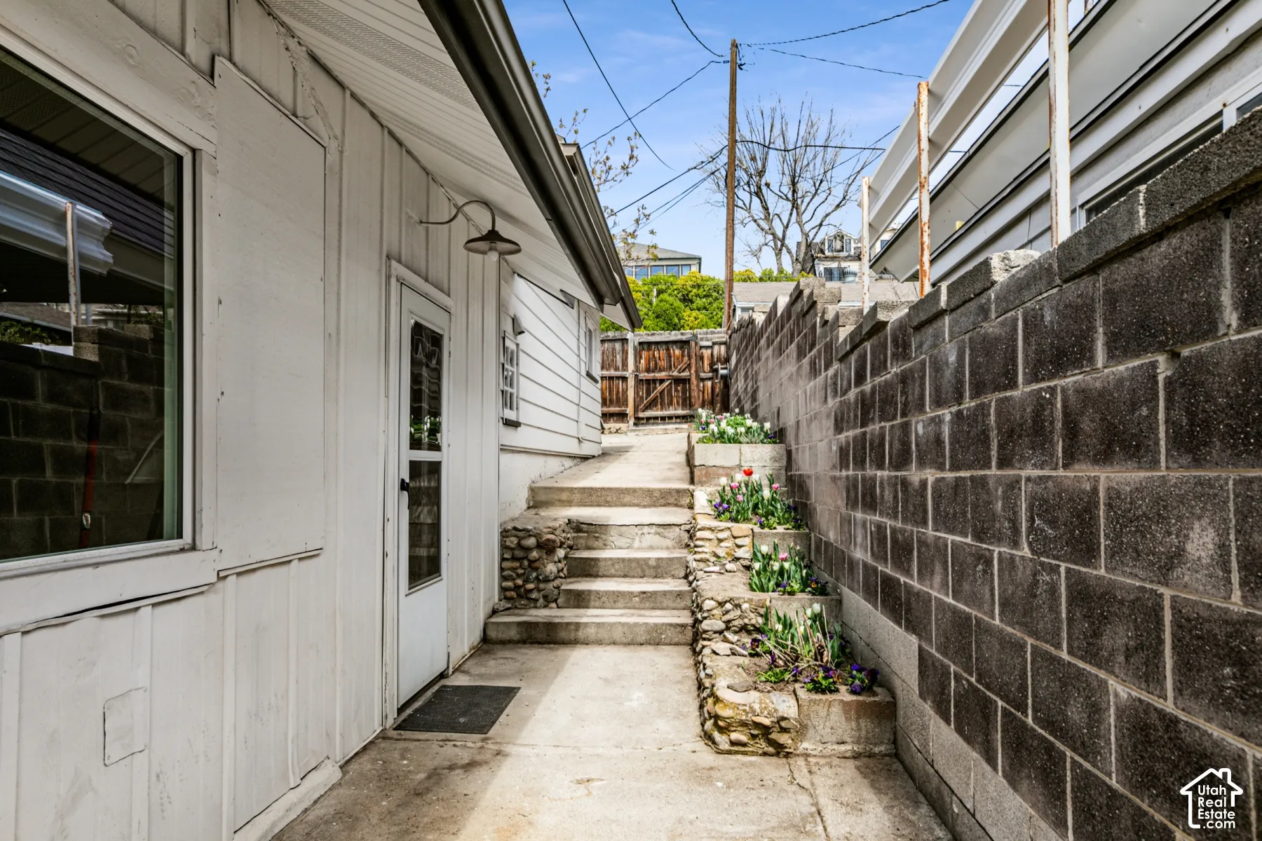 Exterior concrete walkway featuring tiered stone-accented planters, a solid concrete block perimeter wall, and a fixed industrial-style wall-mounted light fixture