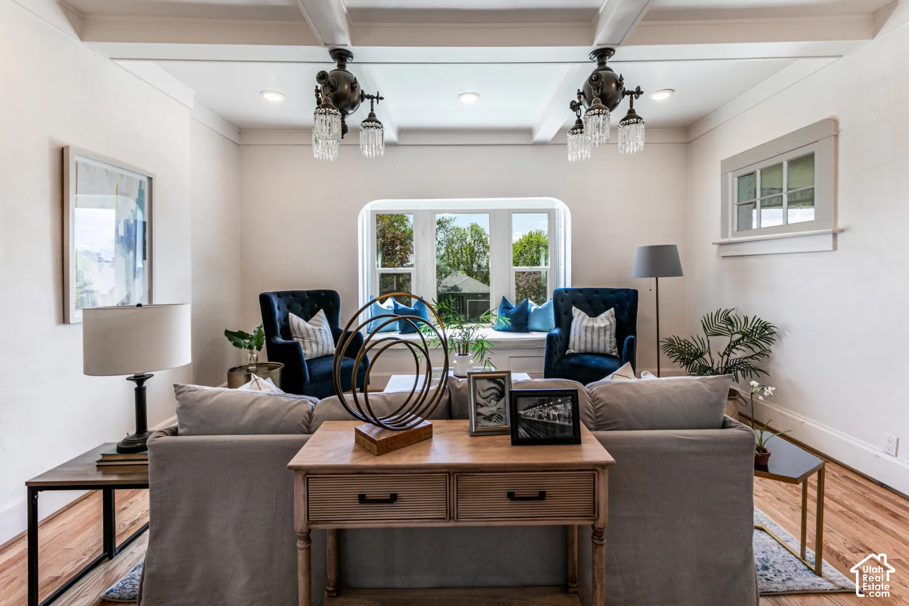 Living space featuring a coffered ceiling, dual multi-arm crystal light fixtures, wood-finish flooring, a deep-set bay window with built-in bench seating, and a multi-pane transom window