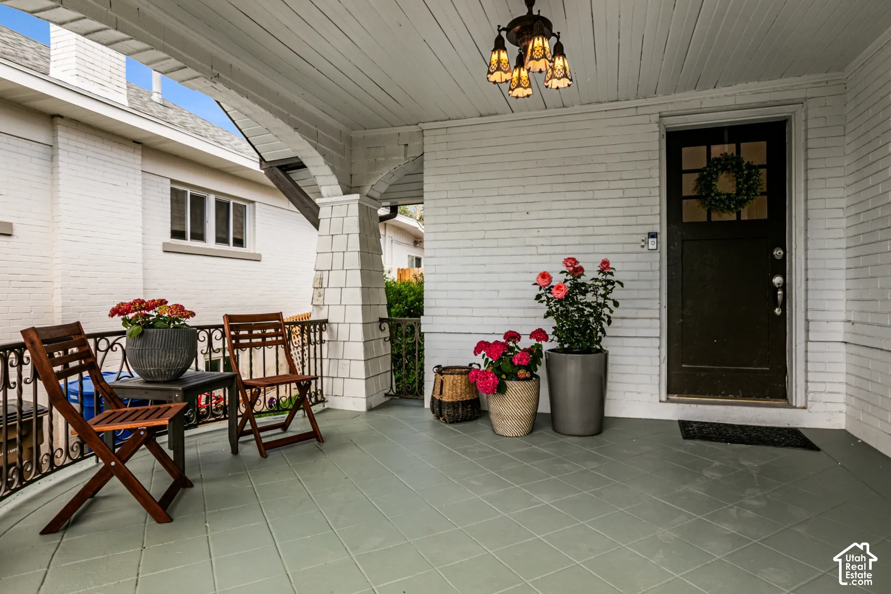 Covered front porch featuring painted brick siding, arched architectural detailing, decorative iron railing, tile flooring, and a multi-light pendant fixture