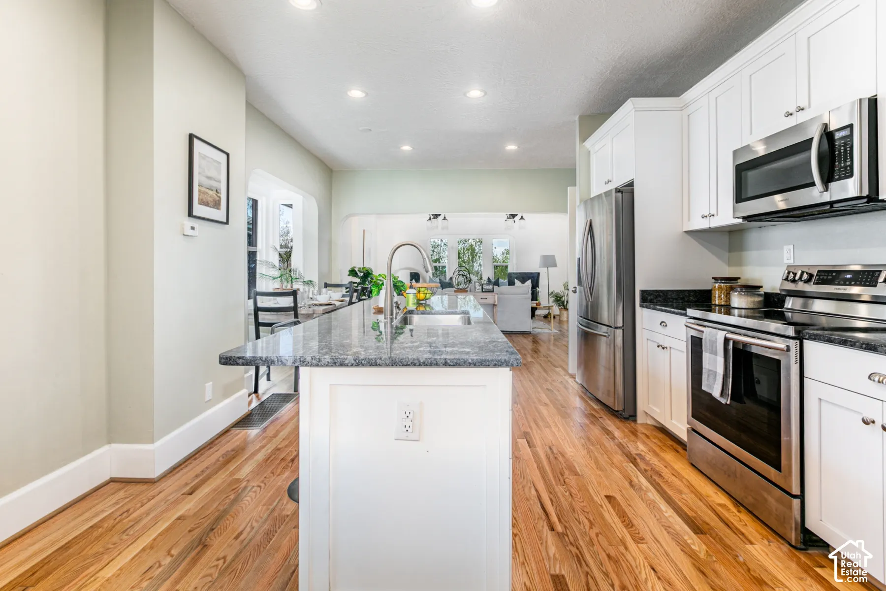 Kitchen featuring white shaker cabinetry, stone countertops, stainless steel appliances, a central island with an undermount sink, and wood-finish flooring