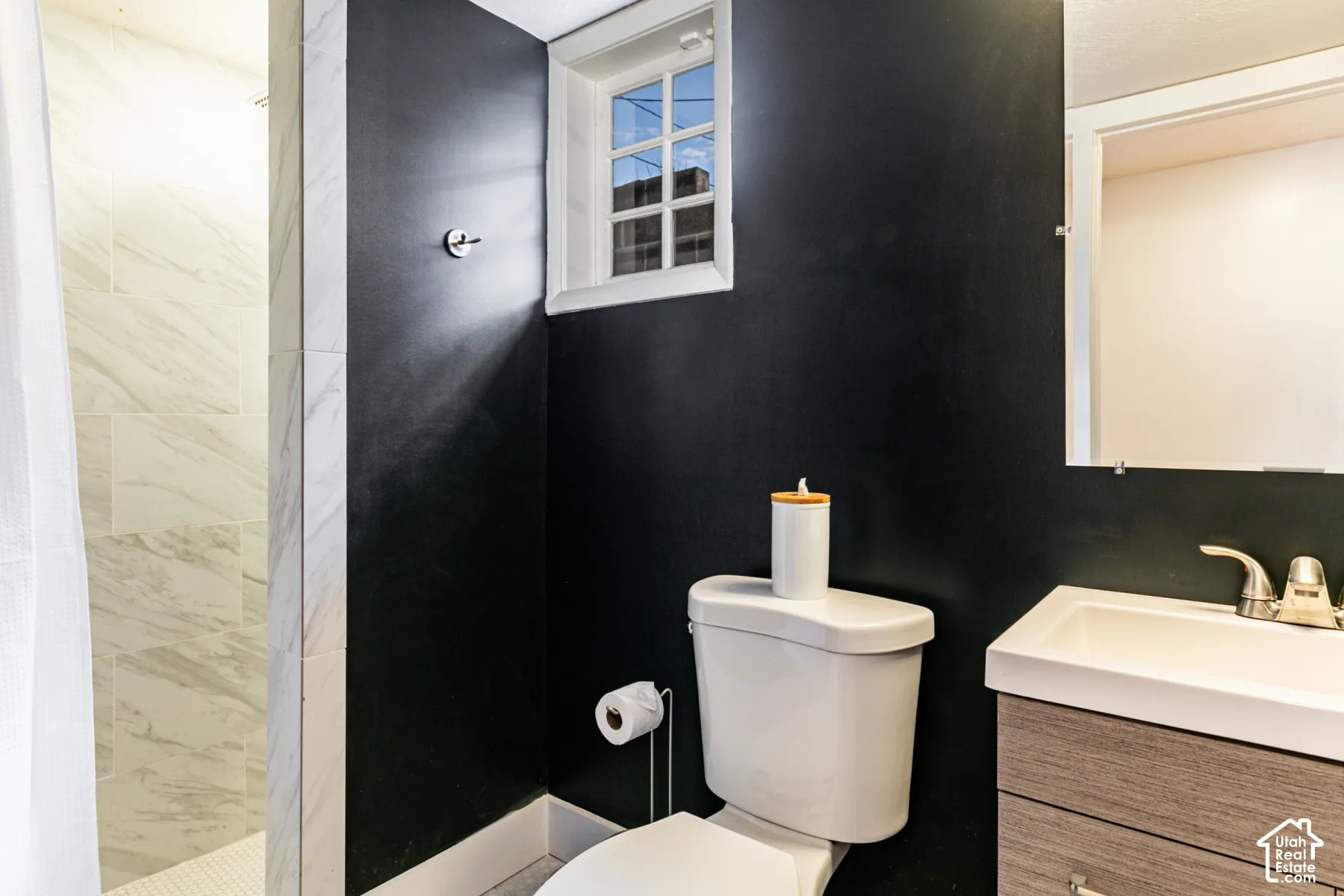 Bathroom featuring a wood-finish vanity with a rectangular sink, wall-mounted mirror, multi-pane window, and walk-in shower with light-toned wall tile