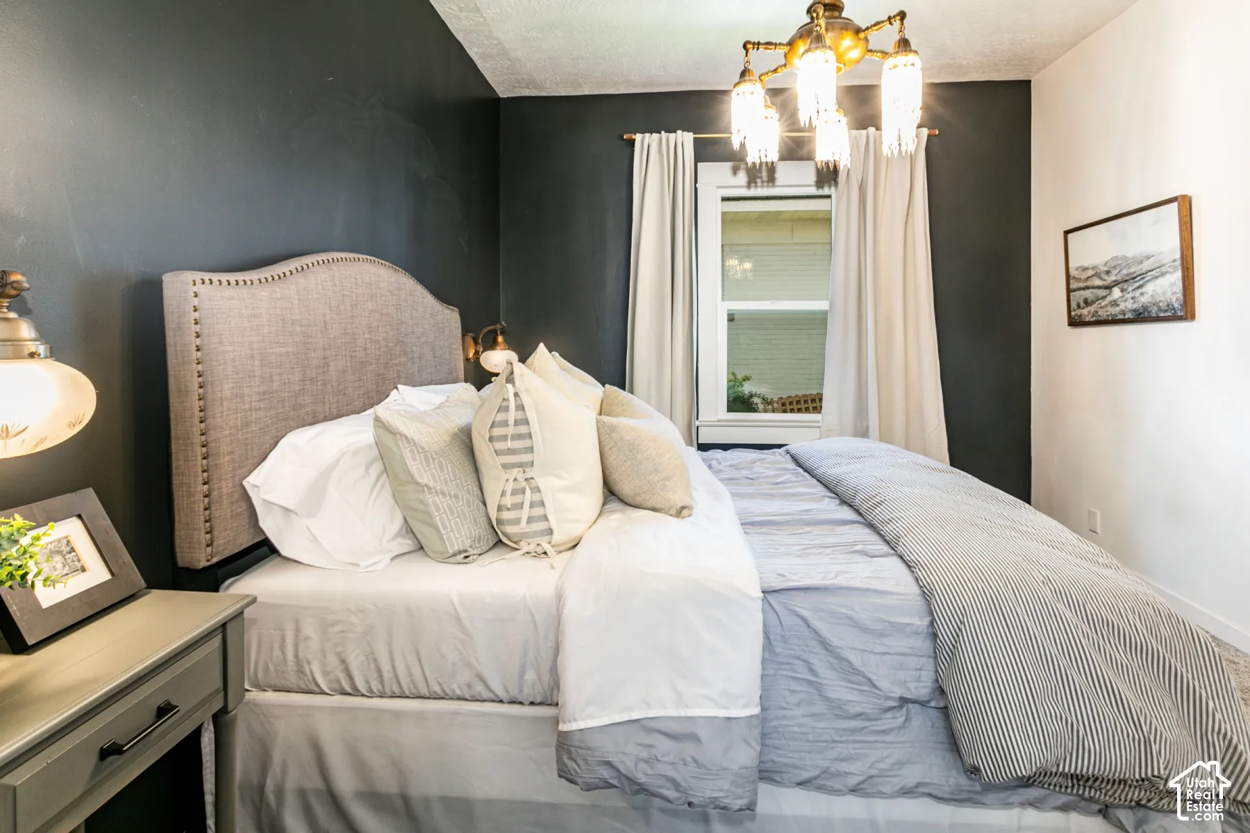 Bedroom featuring a dark-toned accent wall, multi-light brass chandelier, and window with casing trim