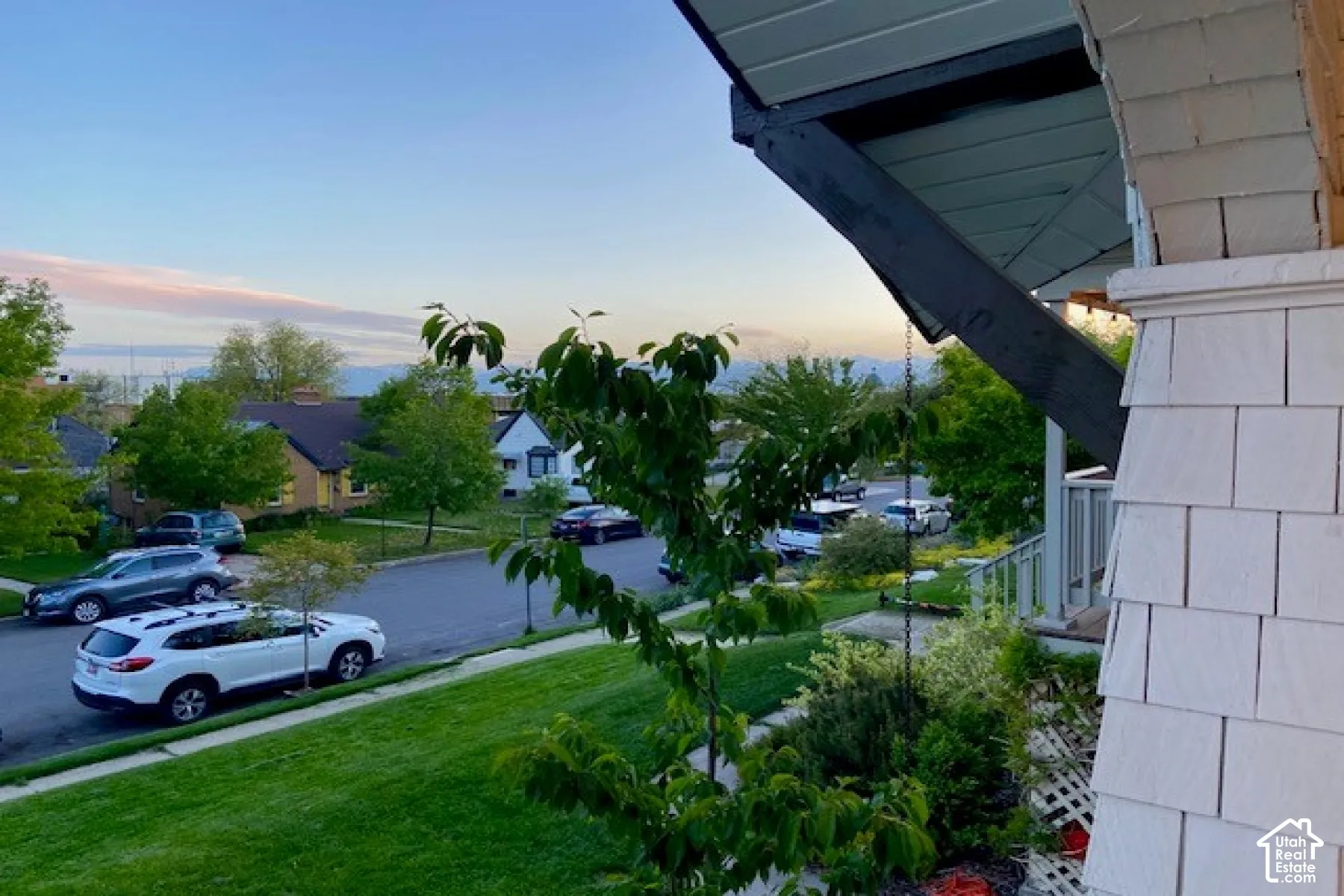 Covered front porch featuring white shake siding and dark-stained structural framing with an exposed rafter tail, overlooking a manicured lawn and a mountain backdrop