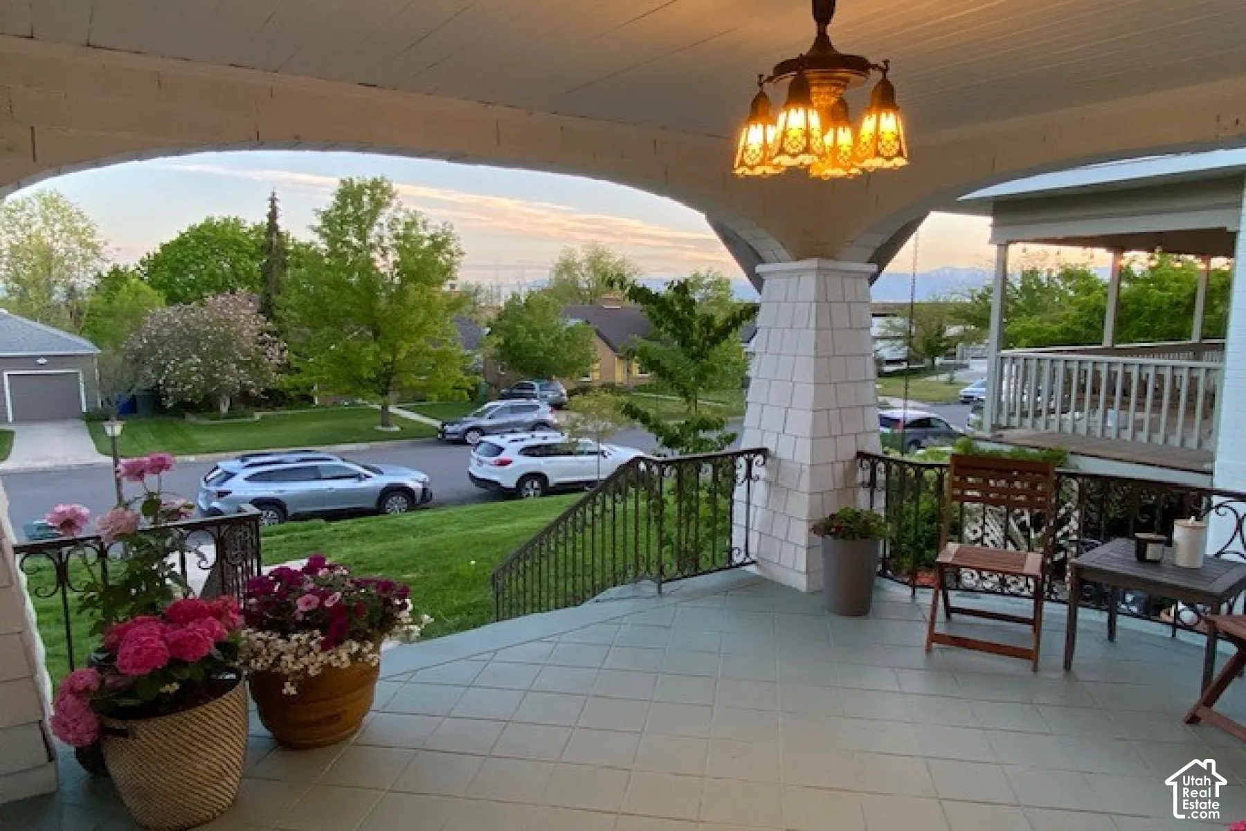 Covered porch featuring tiled flooring, a multi-arm pendant light fixture, shingle-style column cladding, and ornate metal railings