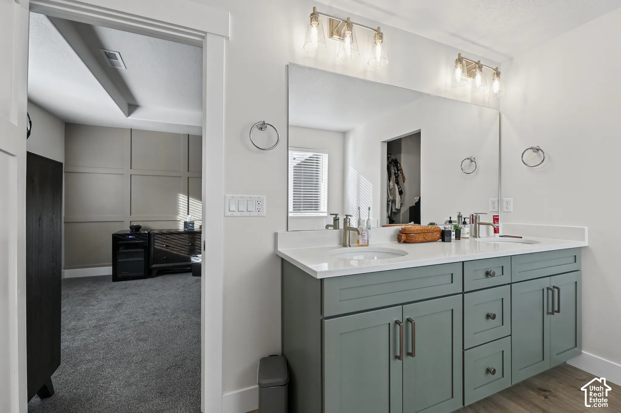 Dual-sink vanity with a solid-surface countertop, cabinetry in a muted blue-green finish, metallic hardware, large rectangular wall mirror, and dual three-light fixtures