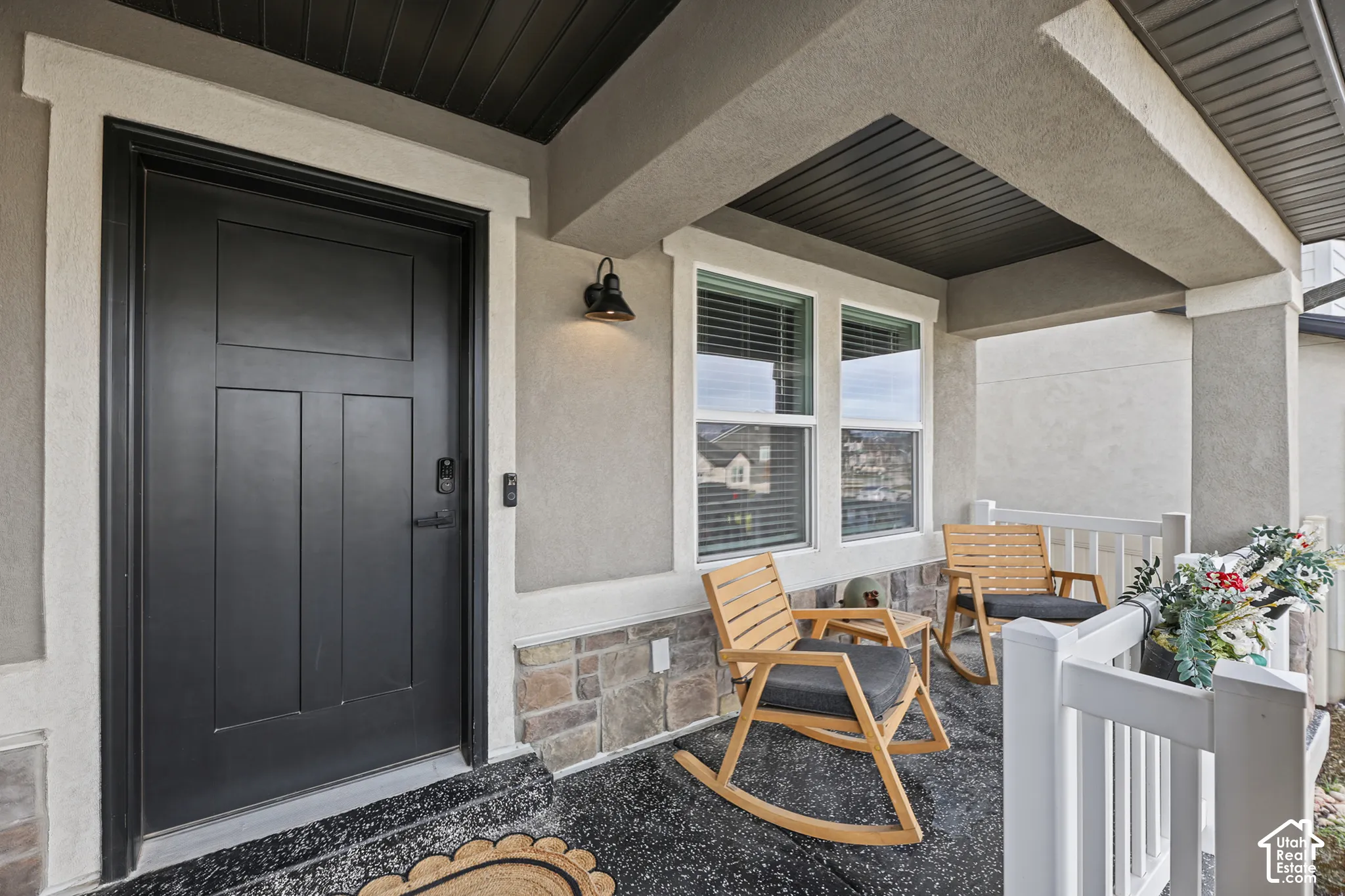 Covered entryway featuring a solid-panel door with matte black hardware, stone veneer wainscoting, white railings, and a wall-mounted exterior light fixture