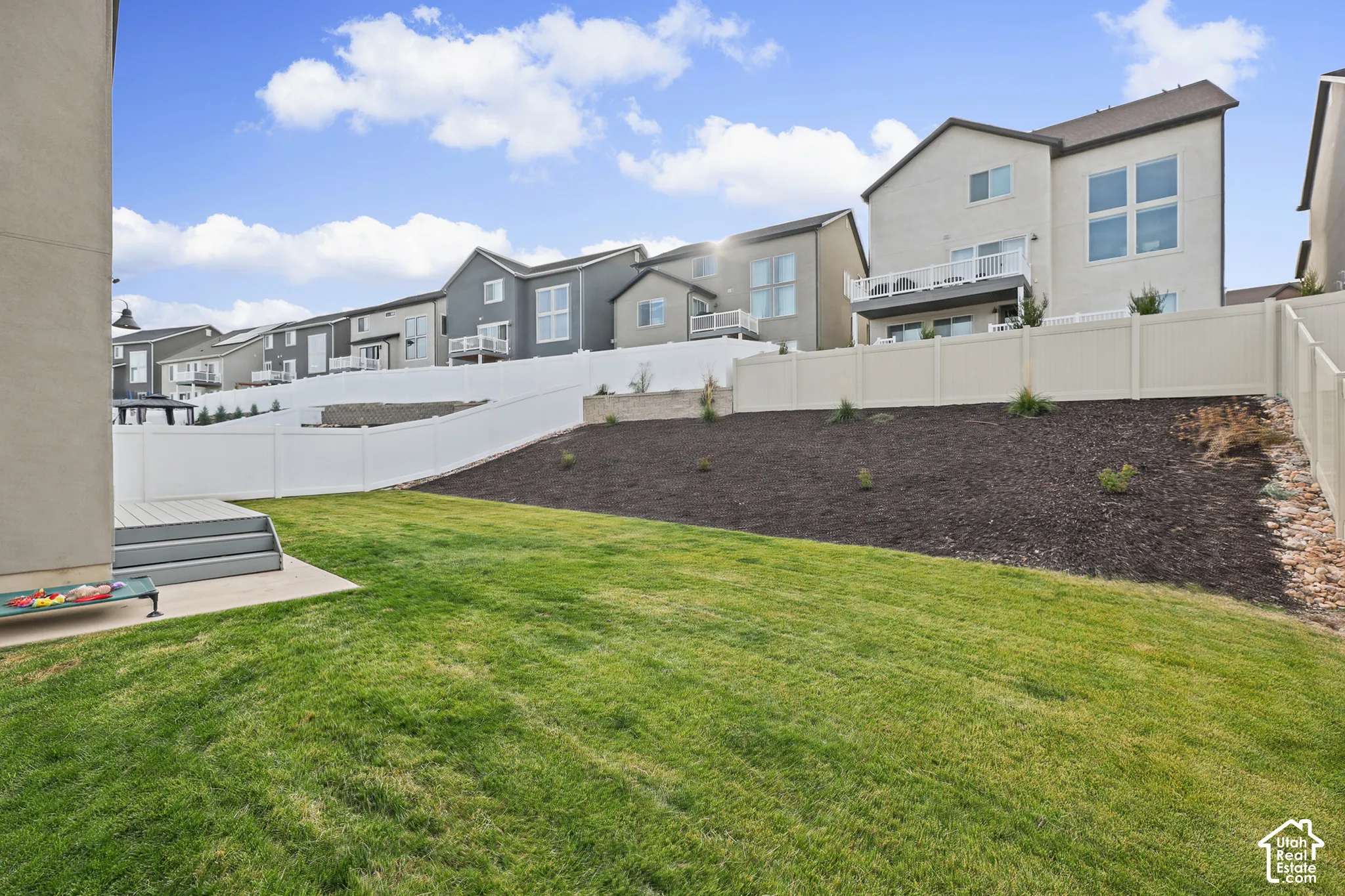 Rear exterior featuring a tiered backyard with a landscaped slope, turf lawn, composite decking, and a white vinyl privacy fence