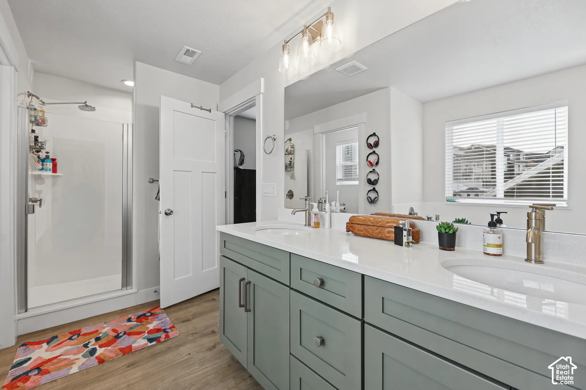 Dual-sink vanity with light-colored solid surface countertops, sage green Shaker-style cabinetry, wood-finish flooring, and a glass-enclosed shower