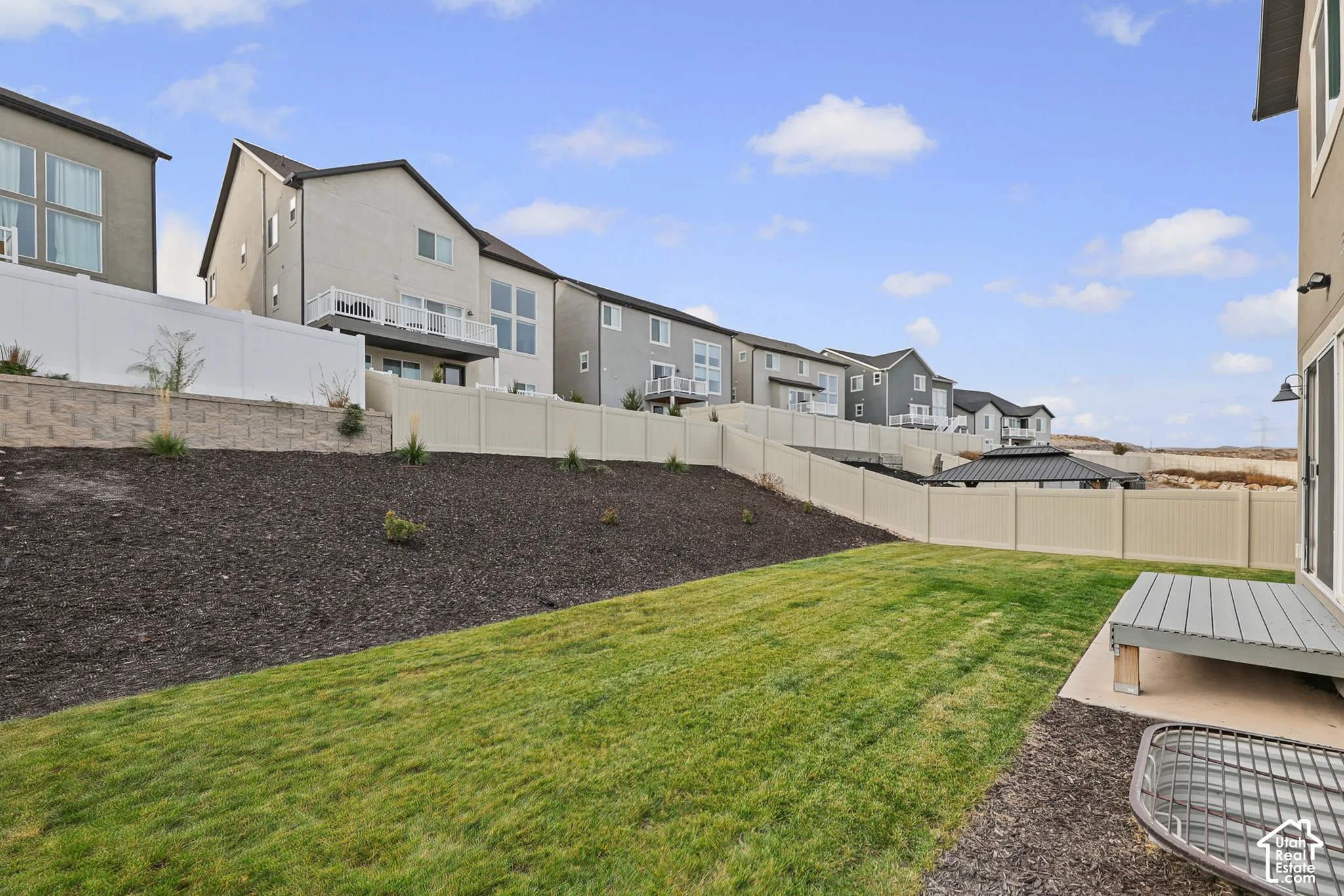 Backyard space featuring a leveled grass lawn, mulched garden beds, a concrete patio, a raised wooden platform, and perimeter vinyl fencing