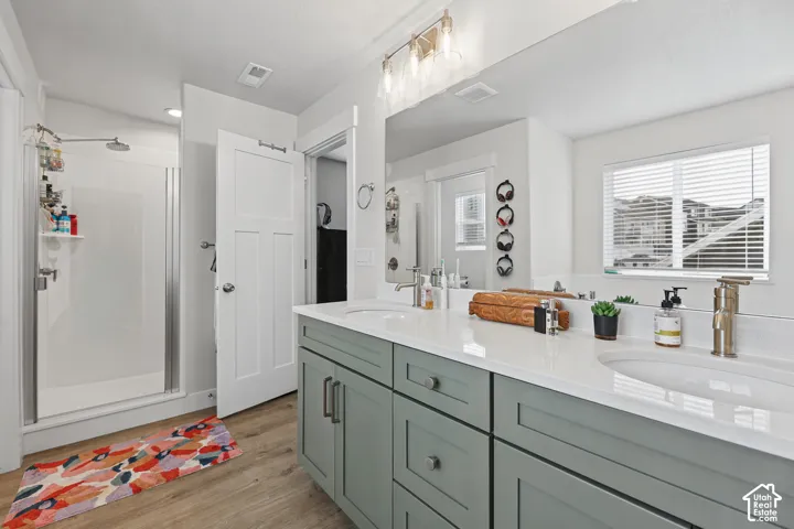 Dual-sink vanity with light-colored solid surface countertops, sage green Shaker-style cabinetry, wood-finish flooring, and a glass-enclosed shower