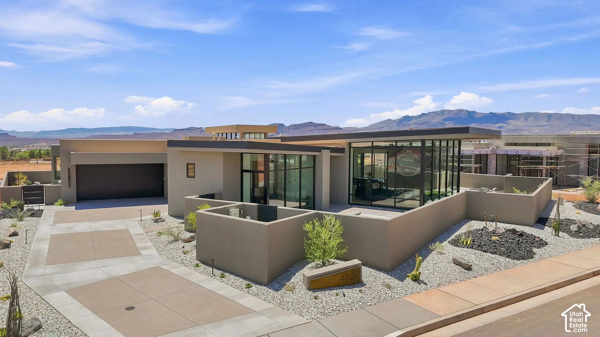Contemporary architectural exterior featuring flat roof lines, floor-to-ceiling glass paneling, concrete driveway, and xeriscaped desert landscaping against a mountain backdrop