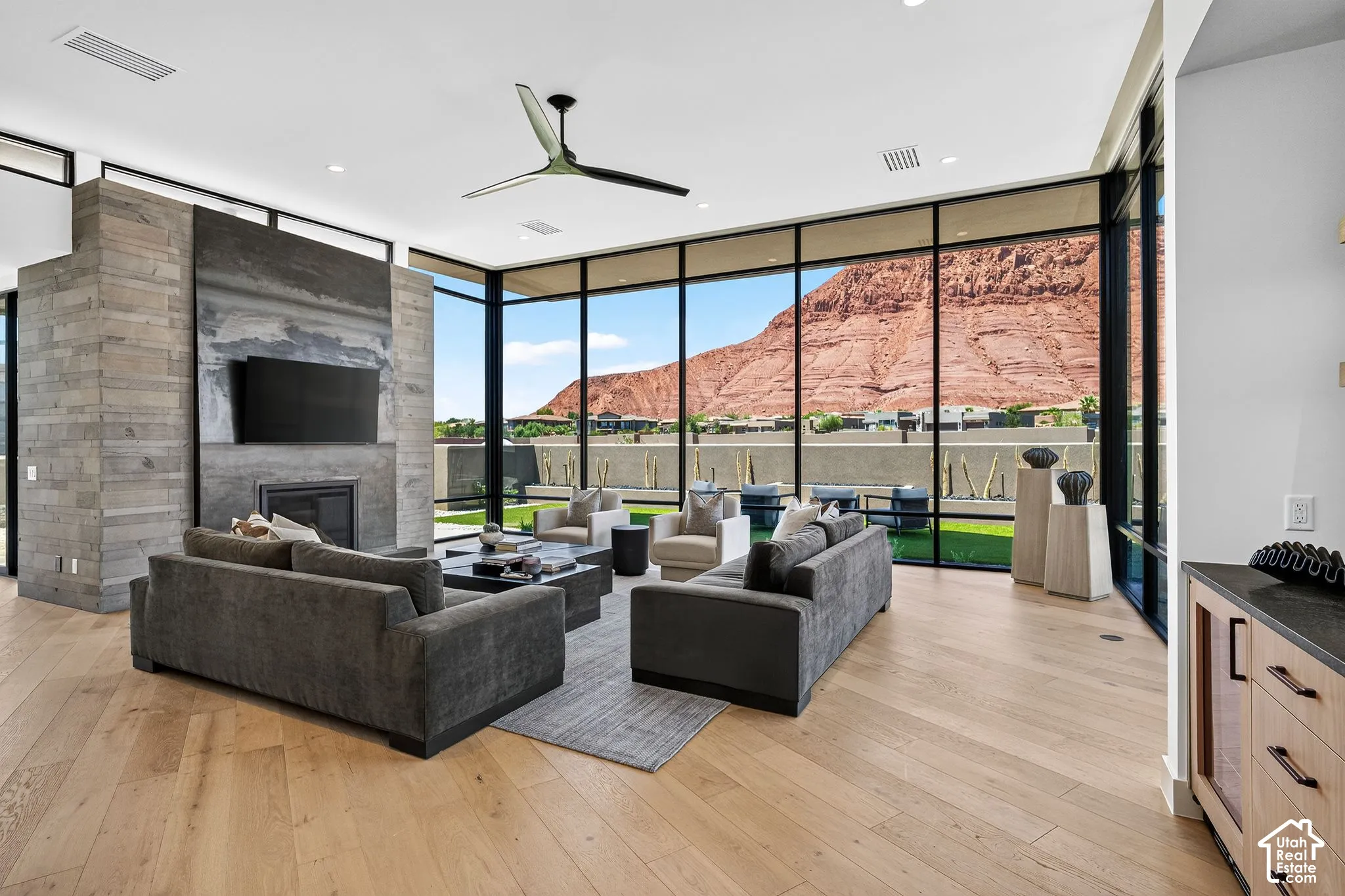 Open-concept interior featuring floor-to-ceiling glass walls, wood-finish flooring, a stone-clad fireplace surround, and prominent mountain scenery