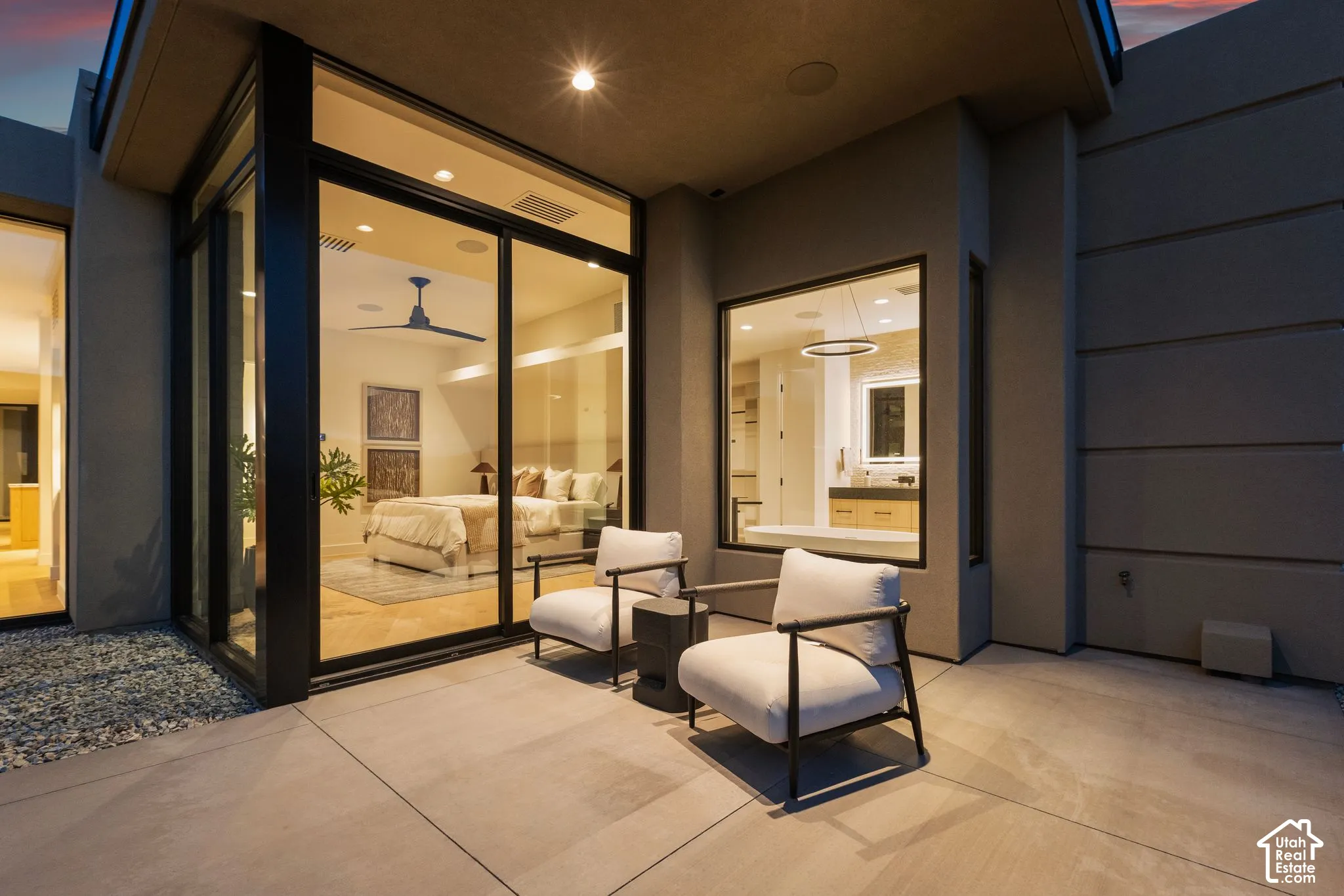 Modern covered patio featuring smooth concrete paving, expansive floor-to-ceiling glass wall panels with black frames, recessed ceiling lighting, and built-in exterior audio speakers