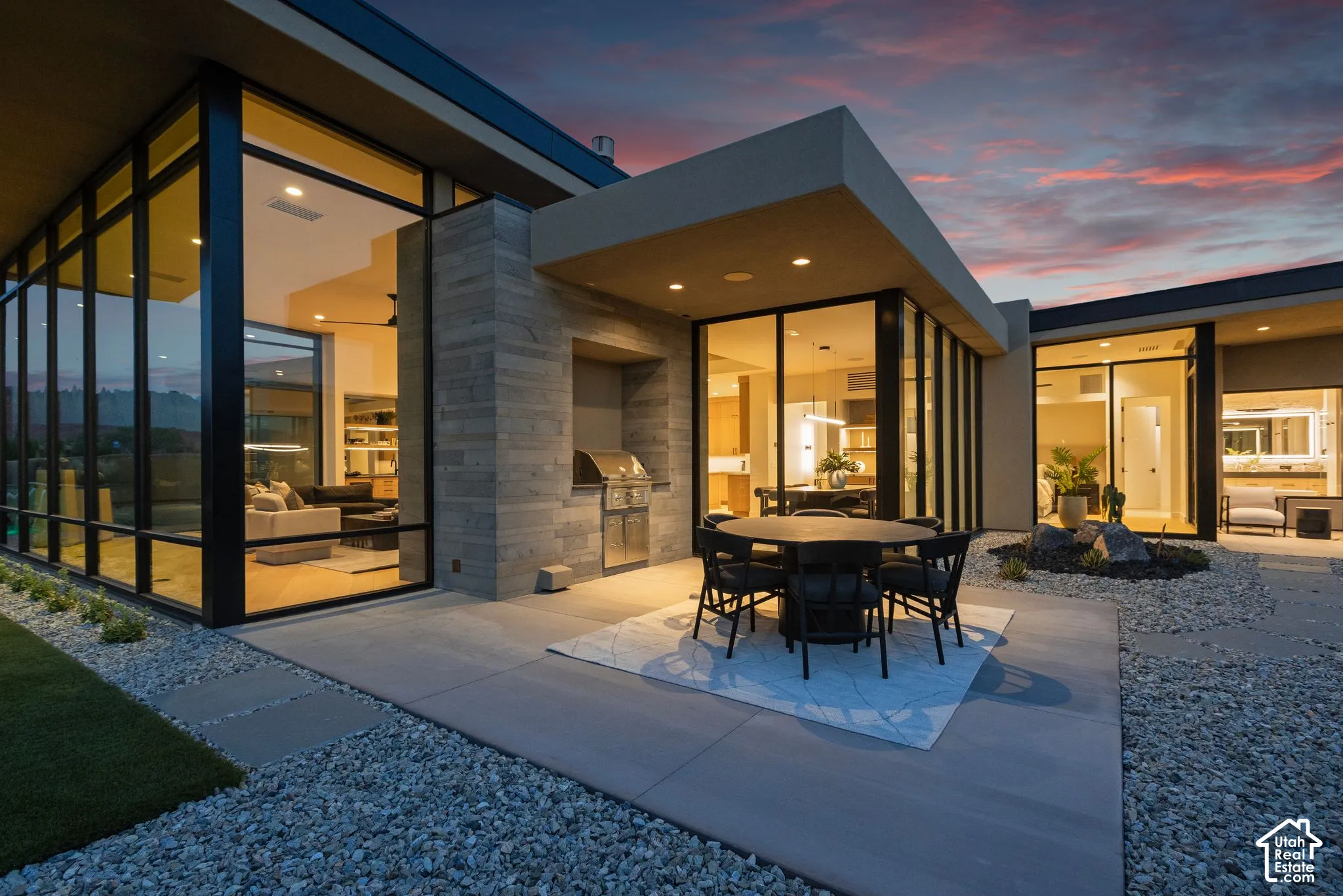 Modern exterior featuring floor-to-ceiling glass windows, integrated stone-clad outdoor kitchen, concrete patio, and decorative gravel landscaping