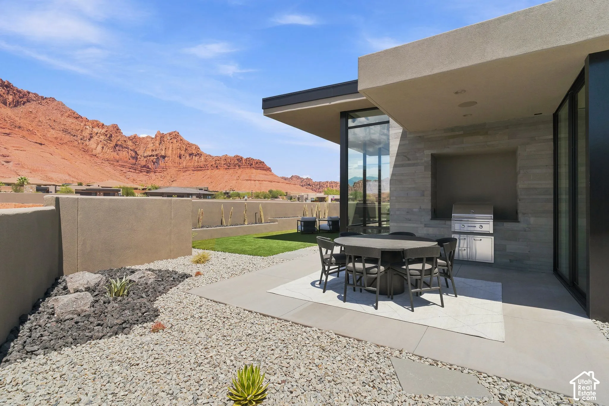 Modern covered patio featuring a built-in stainless steel grill, natural stone veneer siding, floor-to-ceiling glass wall, low-maintenance hardscaping, and prominent red rock mountain backdrop