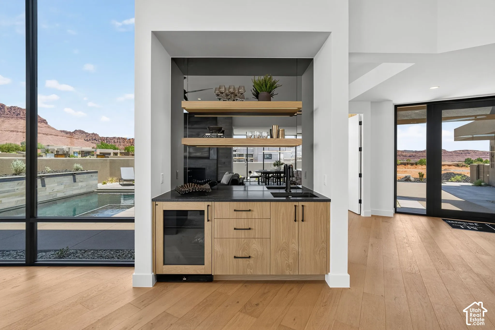 Built-in wet bar featuring light wood cabinetry, matte black hardware, integrated beverage cooler, dark stone-finish countertop, and decorative floating wood shelves against a mirrored backsplash