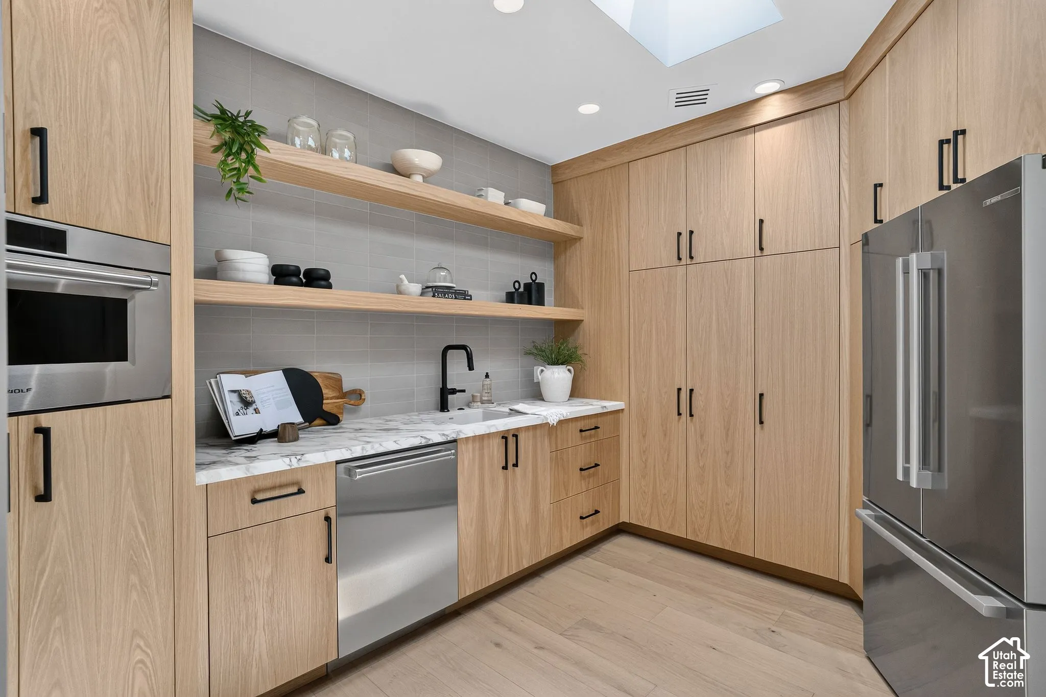 Custom light-wood cabinetry with matte black hardware, stone-surface countertops, built-in stainless steel appliances, stacked tile backsplash, and wood-finish flooring
