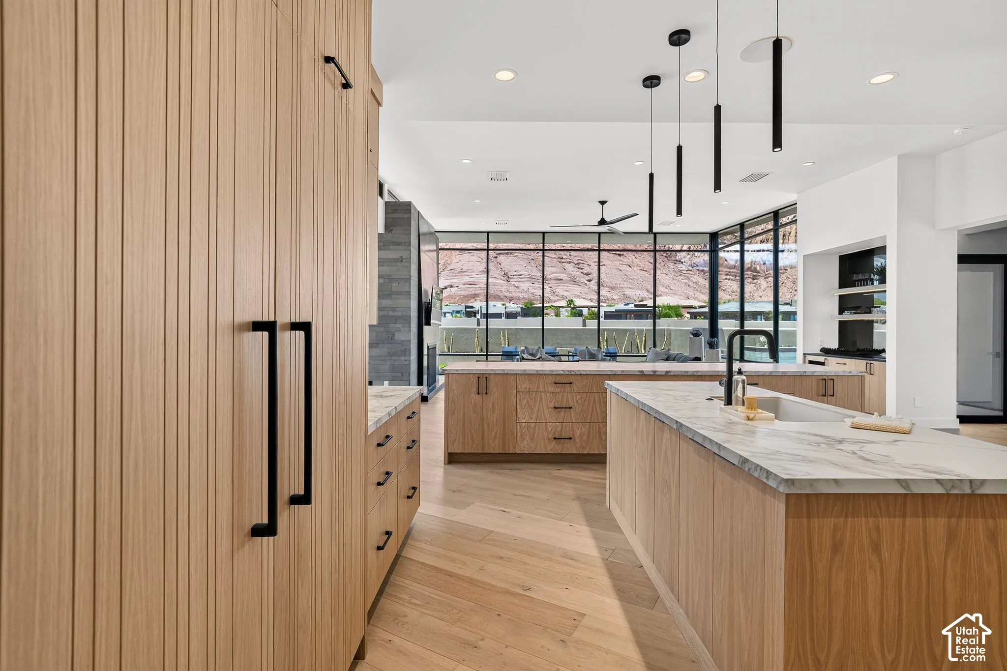 Custom light-oak cabinetry with linear detailing, expansive stone-surface kitchen island, matte black hardware and fixtures, floor-to-ceiling windows with mountain scenery, and light-tone wood-finish flooring