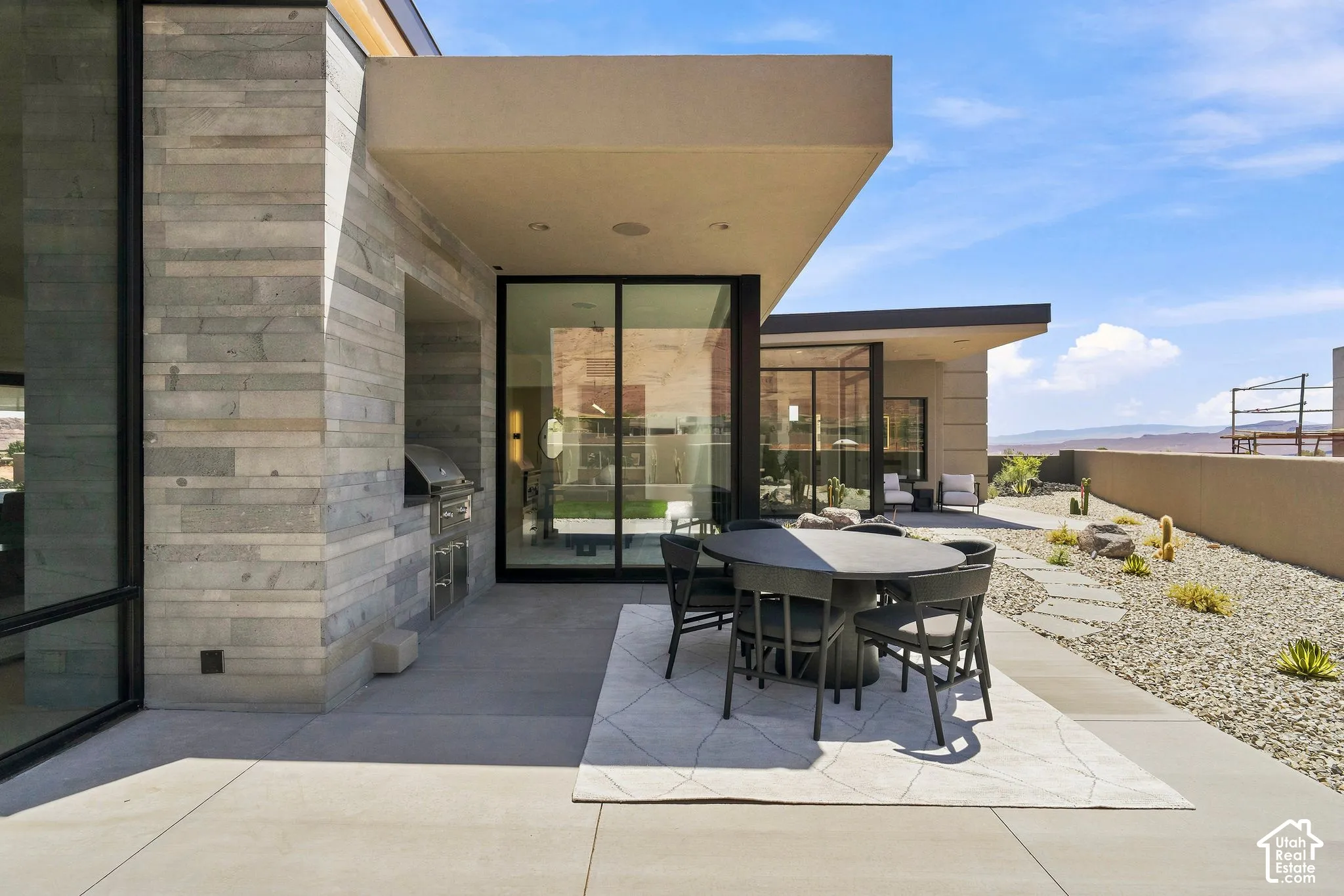 Covered patio featuring linear stone masonry, built-in stainless steel outdoor kitchen, expansive sliding glass wall systems, concrete hardscaping, and xeriscaped desert grounds